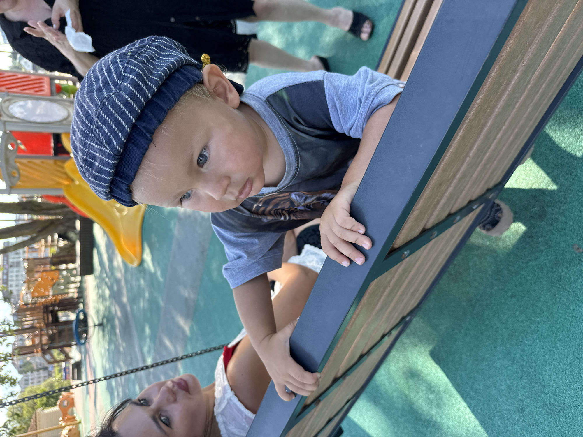 A child wearing a blue pinstripe cap and a grey t-shirt with a tiger graphic is seated on a wooden bench, holding onto the dark blue railing. The child's face is turned to the left, with blue eyes and light-colored hair. To the left, a woman with dark hair and red lipstick is partially visible, also seated on the bench. In the background, a playground with colorful structures and slides is visible, suggesting an outdoor recreational area. The ground is covered with a green, textured surface. The overall scene appears to be a sunny day at a park or playground, likely in an urban setting, with no specific indication of the city of Sochi, Russia.