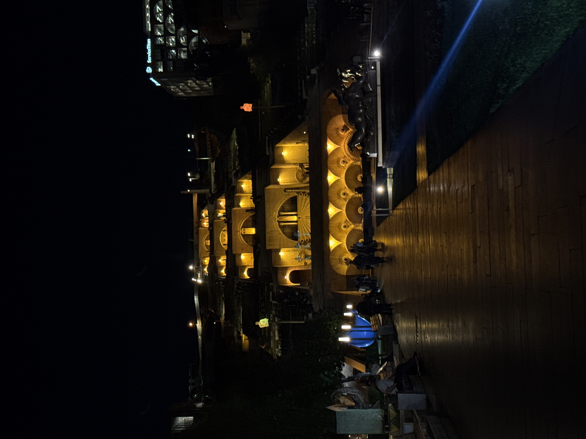 Here is a description of the image:
The image depicts the Cascade monument in Yerevan, Armenia, at night. The structure is illuminated with warm yellow lights highlighting the stepped terraces and architectural details. Several people are visible walking along the wooden pathway adjacent to the monument, with some gathered near the bottom. A blue sculpture stands out against the dark background. A building with brightly lit windows, displaying the name "ServiceTitan," is visible in the upper left corner. The overall environment is dark, with the lights from the Cascade and surrounding buildings providing the primary illumination.