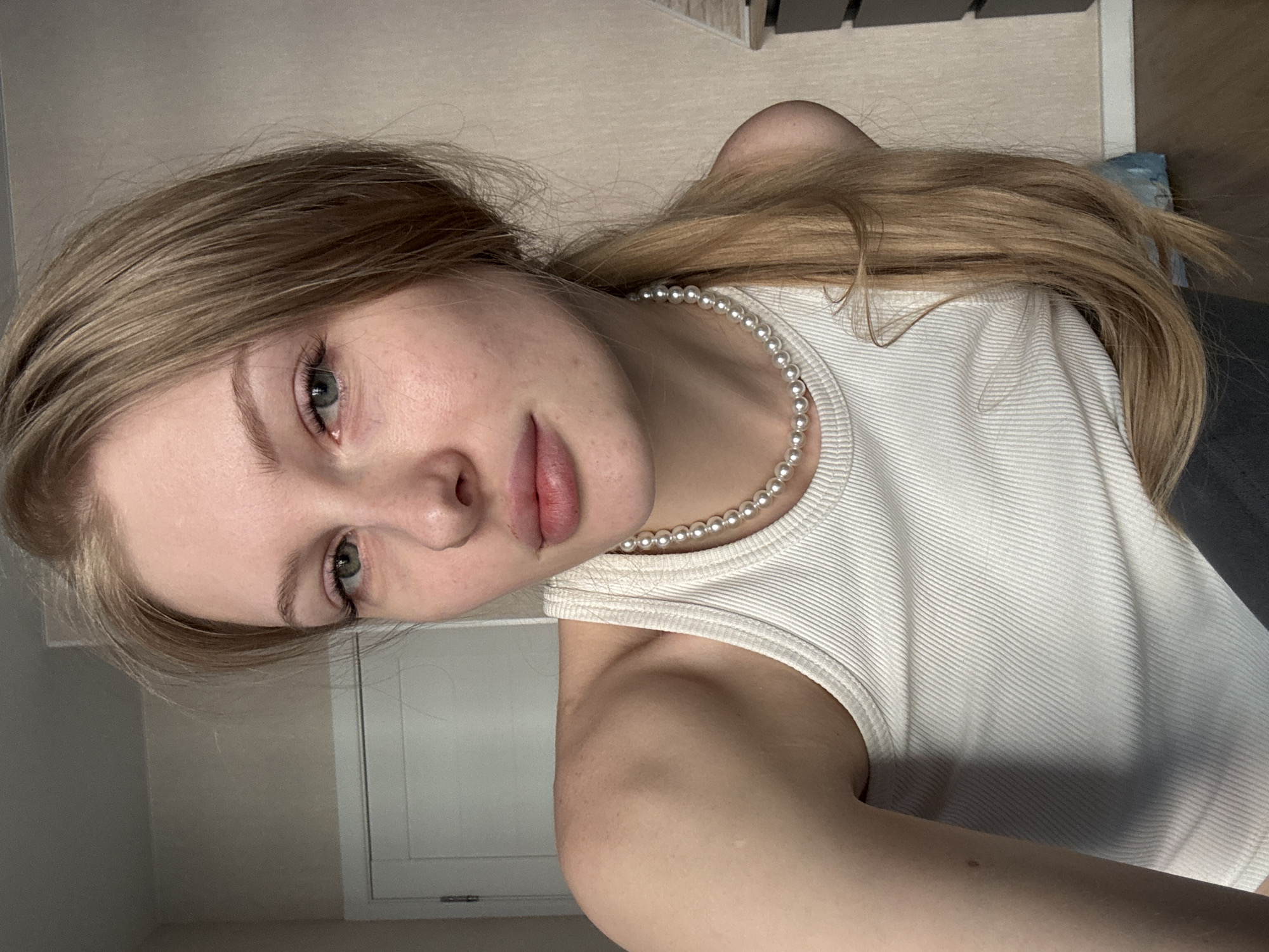 A young woman, appearing to be in her late teens or early twenties, is depicted in a close-up selfie portrait, likely taken indoors in Perm, Russia. She has long, straight light blonde or light brown hair, parted on the side and flowing over her right shoulder. Her eyes are a light green or grey, and she has full, naturally pink lips. She looks directly at the camera with a calm and neutral expression.
She is wearing a simple white ribbed tank top, and a pearl necklace is visible around her neck, featuring uniformly sized white spherical pearls. The background is minimalist, consisting of a light beige or off-white wall, suggesting a domestic setting such as a room in a home. Towards the upper right of the frame, a darker, indistinct area hints at other room features or furniture. The lighting is soft and even, consistent with indoor ambient light. No text is visible in the image.