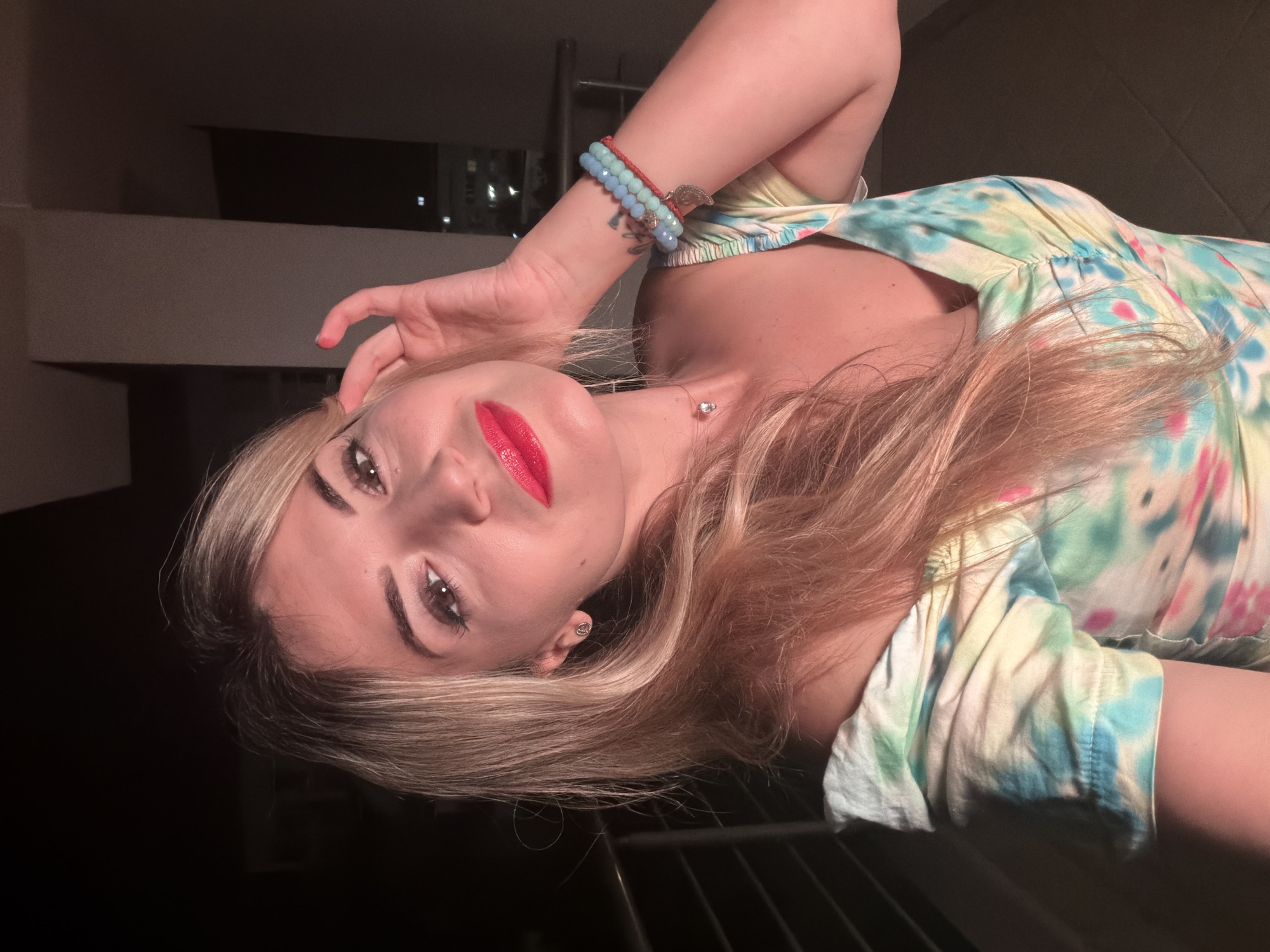 A medium shot captured at night from a balcony in Budva, Montenegro, shows a young woman with blonde hair and bright red lipstick. She is looking at the camera, her right hand touching her hair near her temple. She is wearing an off-the-shoulder dress with a tie-dye pattern in shades of green, yellow, blue, and pink. A necklace with a single clear stone is visible on her neck, and on her right wrist, she wears three beaded bracelets with a small heart-shaped charm. The background is dark, with a railing and some blurred lights suggesting an urban setting.