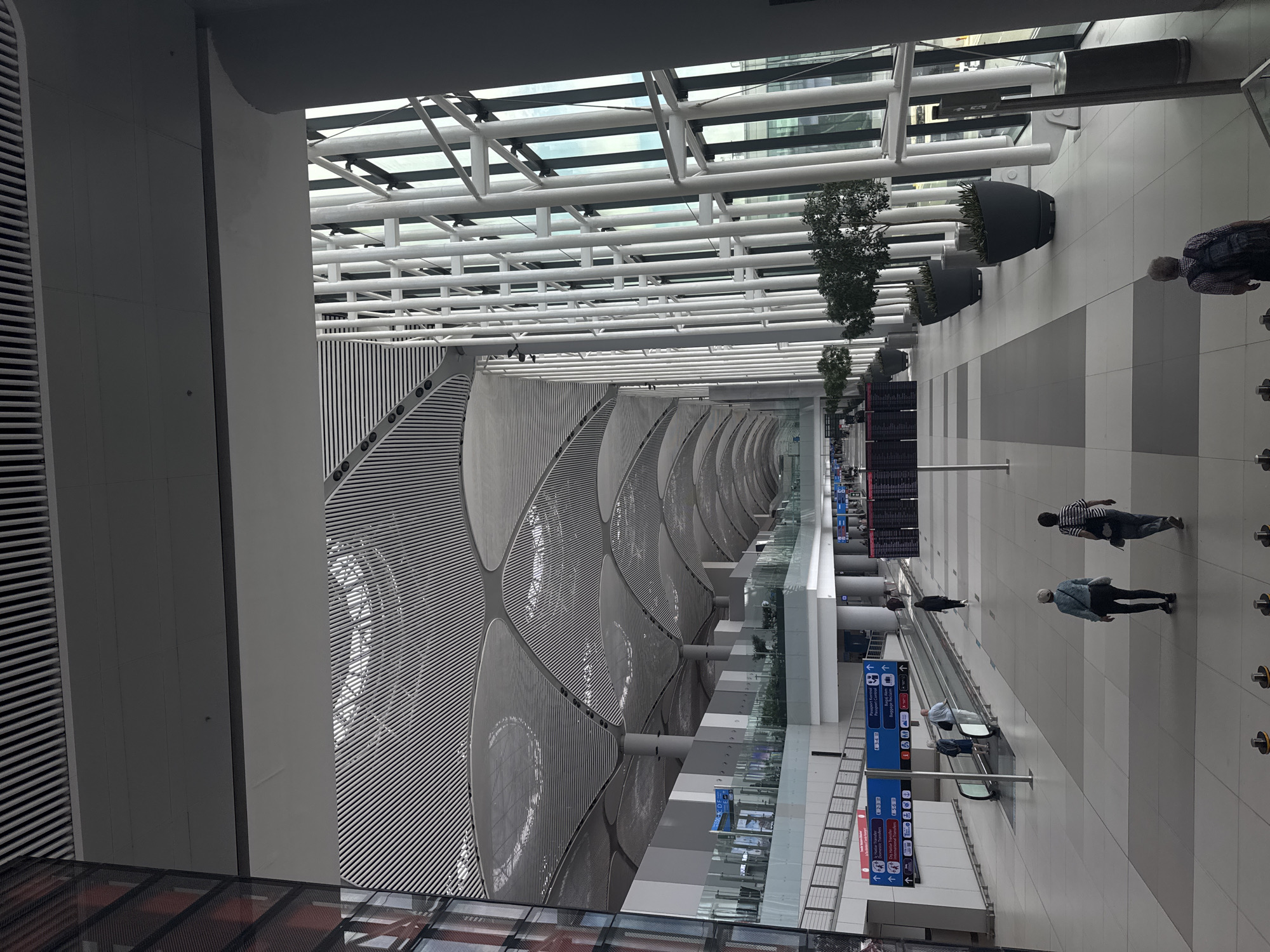 This image captures the interior of a modern airport terminal, specifically located in Arnavutköy, Turkey, which is the site of the new Istanbul Airport (IST). The vast space is characterized by a high, undulating ceiling with a ribbed, metallic design supported by large, grey columns. To the right, a wall of floor-to-ceiling windows is framed by white, vertical support beams, allowing natural light to flood the terminal and offering a glimpse of the exterior.
The terminal floor is tiled in a pattern of light and dark grey squares. A moving walkway, or travelator, runs diagonally across the mid-ground, with several people utilizing it. To the left of the travelator, a series of blue and red signs with directional arrows and text in Turkish and English provide information on passenger transfers and passport control.
In the foreground, three individuals are walking away from the camera. Two women are walking side-by-side, and a man with a backpack is slightly behind them. Further into the terminal, other passengers can be seen walking, standing, and using the moving walkway. Large, dark planters containing green foliage are placed along the right side of the terminal, accentuating the contemporary design. In the distance, departure boards display flight information. The overall atmosphere is one of organized transit within a spacious, architecturally striking airport environment.