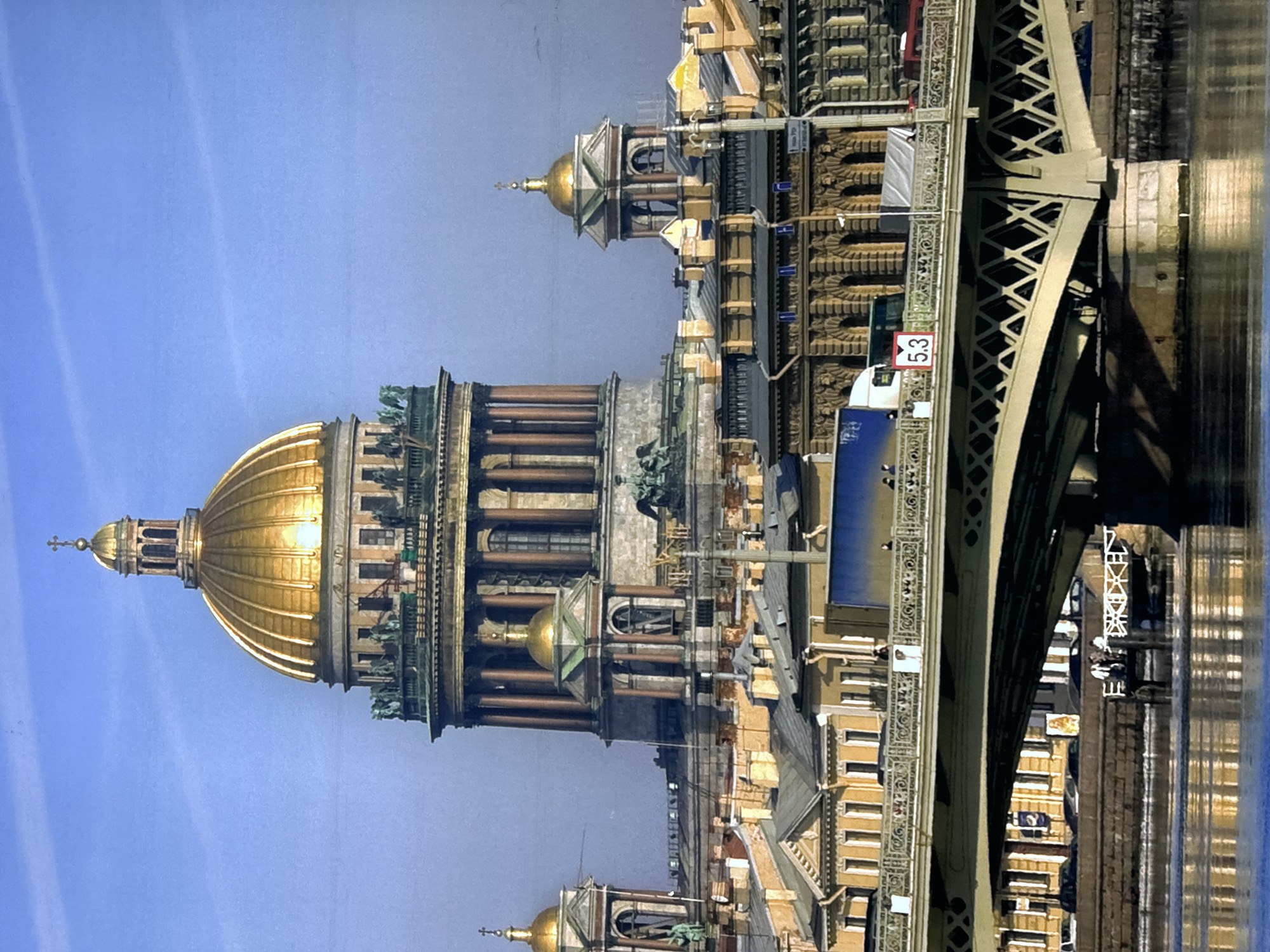 The image shows St. Isaac's Cathedral and a bridge in Saint Petersburg, Russia. The cathedral features a large golden dome and classical architectural details. A bridge with a lattice design passes over what is presumably the Neva River, with reflections of buildings visible in the water. The sky is a clear blue.