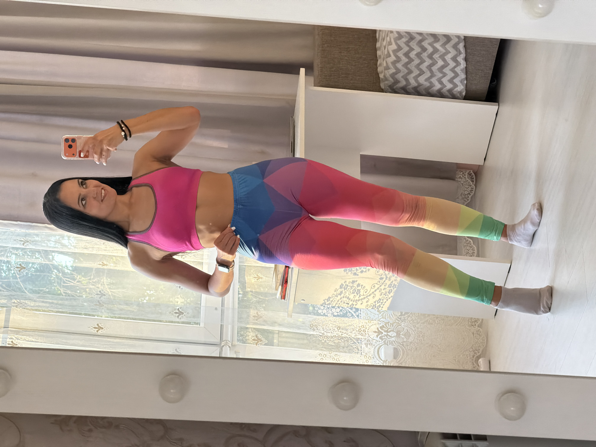 A woman is taking a mirror selfie in a room. She is wearing a pink sports bra and colorful leggings with a rainbow pattern. She is also wearing white socks. The room appears to be a bedroom or dressing room. There is a large mirror in front of her, a window with curtains behind her, a desk with books on it, and a patterned pouf. The overall lighting suggests it is daytime. The image does not contain any information indicating a location in Sochi, Russia.