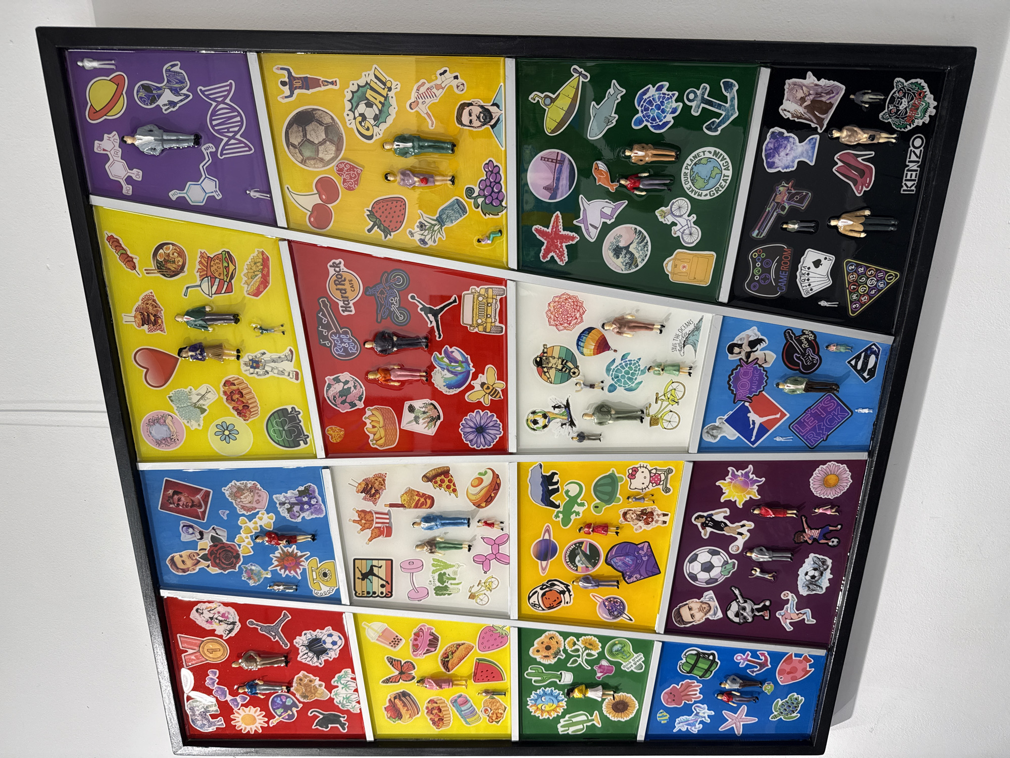 A framed artwork featuring a grid-like pattern filled with stickers and small figurines is displayed on a white wall. The artwork is composed of various colored sections, each containing a collection of stickers representing different themes such as space, sports, food, travel, and pop culture. Miniature figurines, mostly depicting people in various attire, are scattered among the stickers. The frame is black and the artwork has a glossy finish. The scene is indoors, illuminated by ambient light. The artwork seems to be the focal point. There is no information on the location of the city of Moscow, Russia in this image.