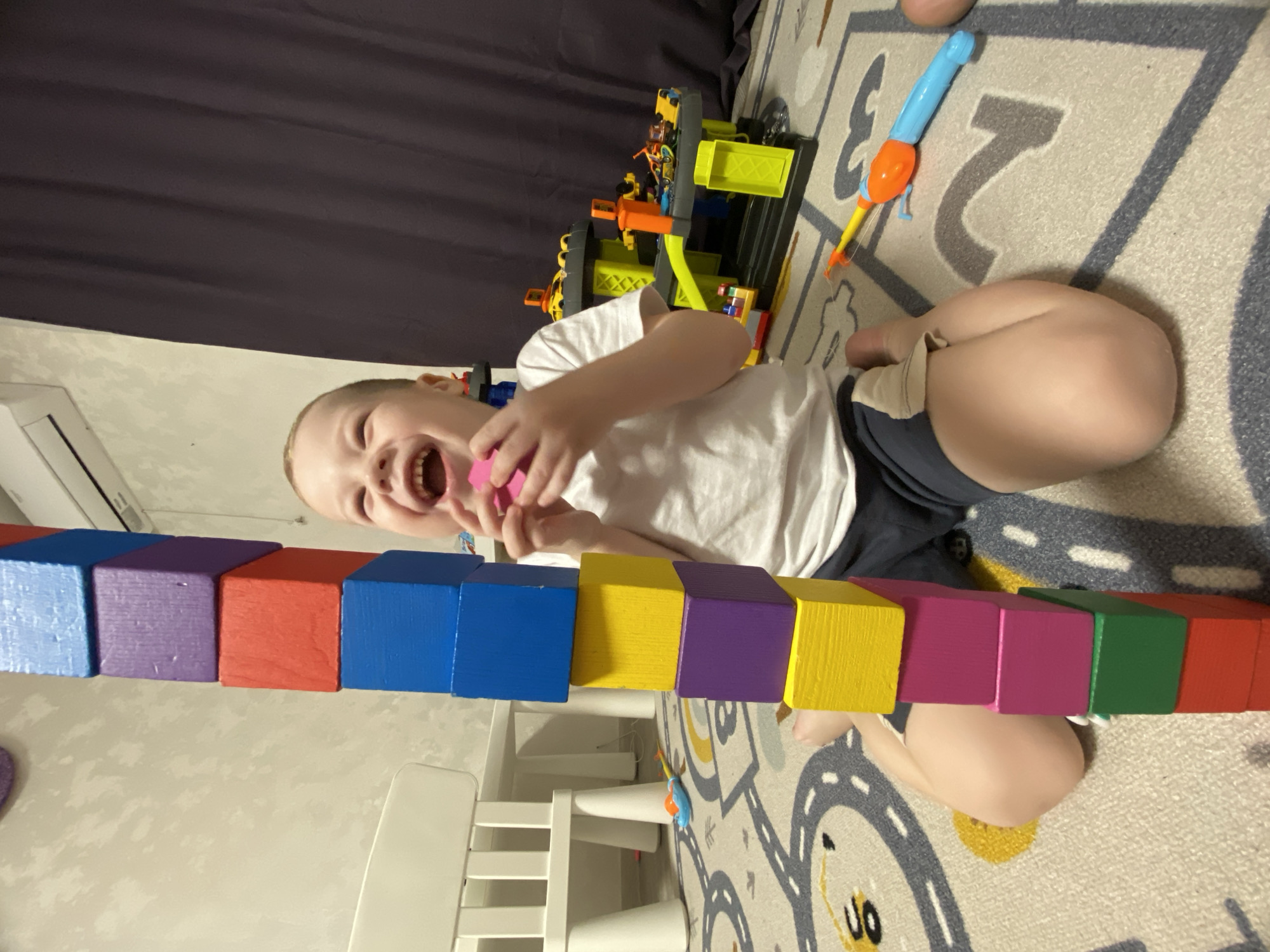A child sits on a patterned rug indoors, laughing and holding a pink object. A line of colorful blocks stretches across the lower portion of the frame, resting on the rug and a white chair/table. Behind the child are several toys and a dark curtain. The location is not evident from the image.