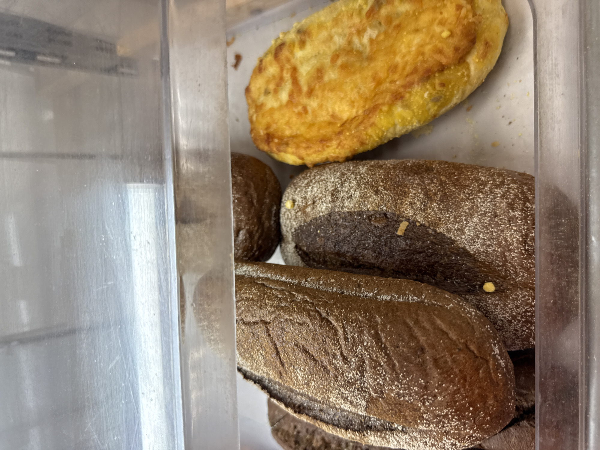 The image shows a display case containing baked goods, likely in a bakery or food establishment in Yekaterinburg, Russia. In the foreground, there are several loaves of dark-colored bread, dusted with flour. To the right, there is a golden-brown pastry or bread item with visible fillings, possibly cheese or vegetables. The display case has a clear plastic front and a metallic shelf above, with some reflections visible on the plastic. The overall lighting suggests an indoor setting.
