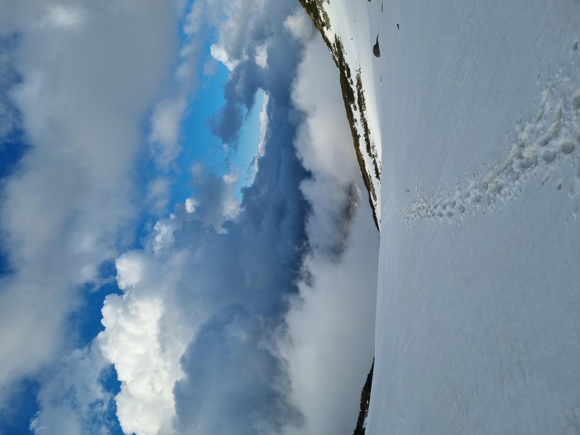 The image shows a mountain landscape in Govedartsi, Bulgaria, with a snow-covered slope on the right side and a dramatic sky on the left. The sky is filled with a mix of white, gray, and blue clouds. A line of vegetation, possibly trees or shrubs, separates the snow from a steep drop. Footprints are visible in the snow, indicating human presence. The overall impression is of a high-altitude scene with variable weather conditions.