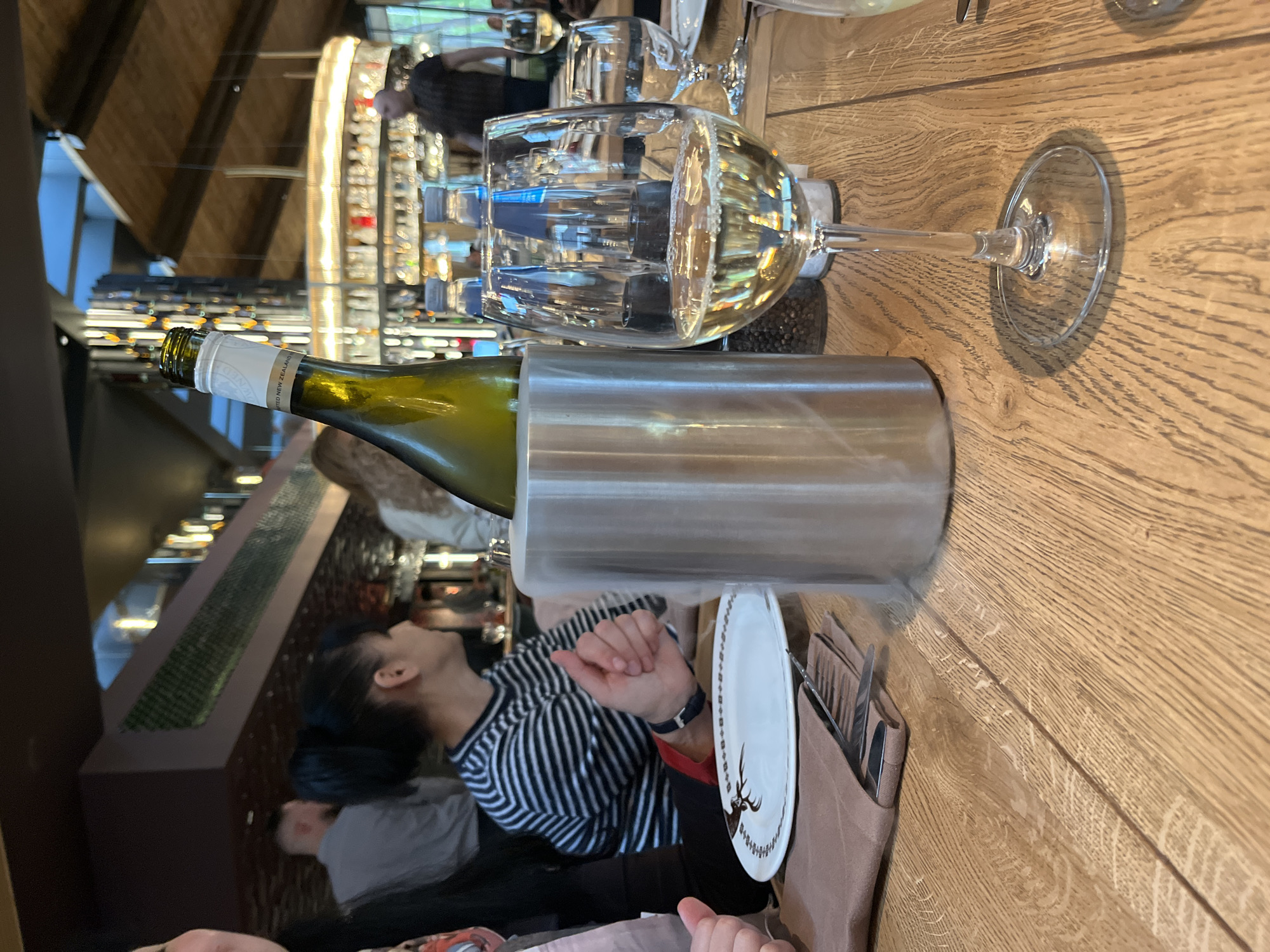 This indoor photograph, taken in Sofia, Bulgaria, captures a tablescape within what appears to be a restaurant or wine bar. The scene is set on a light brown wooden table with visible grain.
A bottle of white wine is prominently displayed, resting within a tall, silver-colored wine cooler. The label on the wine bottle is partially visible, clearly showing "NEW ZEALAND," indicating the wine's country of origin. Adjacent to the wine cooler, a clear stemmed wine glass, partially filled with white wine, stands on the table.
Further back on the table, a white ceramic plate is visible, featuring a distinct brown design, possibly a deer or stag head. Beside the plate, a brown fabric holder contains cutlery, including a fork and knife, suggesting a dining setup.
Several individuals are present in the scene. A person is clearly visible in the mid-ground, wearing a black and white striped long-sleeved top, seated at the table. Their dark hair is tied back, and their arm and hand are visible near the plate. Their gaze appears to be directed upwards. Other blurred figures can be seen in the background, including another person partially visible in the lower left corner and indistinct patrons interacting with a large, illuminated wine display.
The background reveals extensive shelving filled with numerous bottles, likely an impressive wine collection, illuminated by warm lights. This intricate display creates a sophisticated and inviting ambiance. The overall lighting suggests an evening setting within the establishment. No specific emotions are discernible from the visible individuals.
