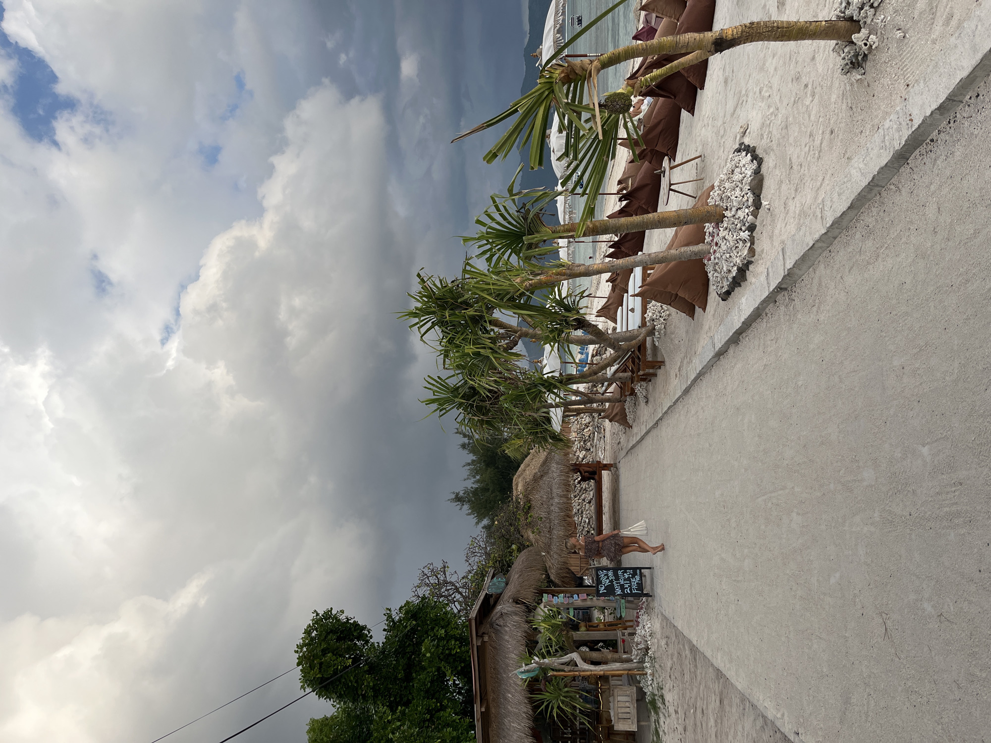 A medium-shot depicts a path on a beach in an unknown location in Indonesia, flanked by a building with a thatched roof on the left and lounge chairs with brown cushions under trees on the right. The sky above is filled with dark, heavy clouds. A woman in a short dress stands near a blackboard menu in front of the thatched building. The path is made of light-colored sand or concrete and appears to lead towards the beach, where boats are visible in the background.