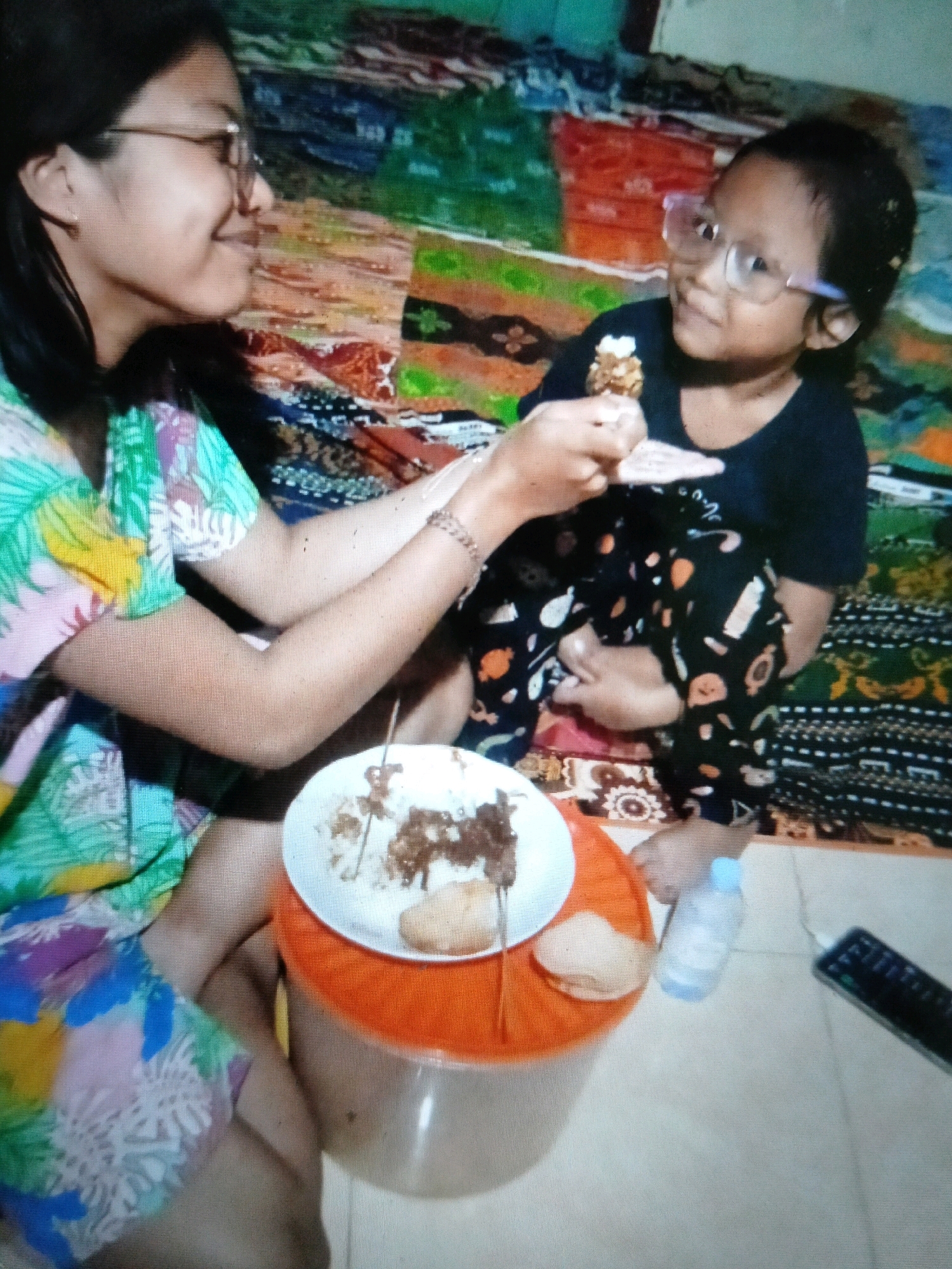 In a room in Puskesmas Keboan, Indonesia, an Indonesian woman wearing glasses and a colorful blouse is feeding a young Indonesian girl, also wearing glasses and a black shirt with a playful pattern on her pants, a piece of food from a plate. The plate contains rice and what appears to be meat skewers and some bread-like items. They are seated on the floor, with a patterned fabric backdrop. To the right of the subjects, a clear plastic water bottle and a black remote control are visible on the floor. The overall impression is a casual mealtime moment.