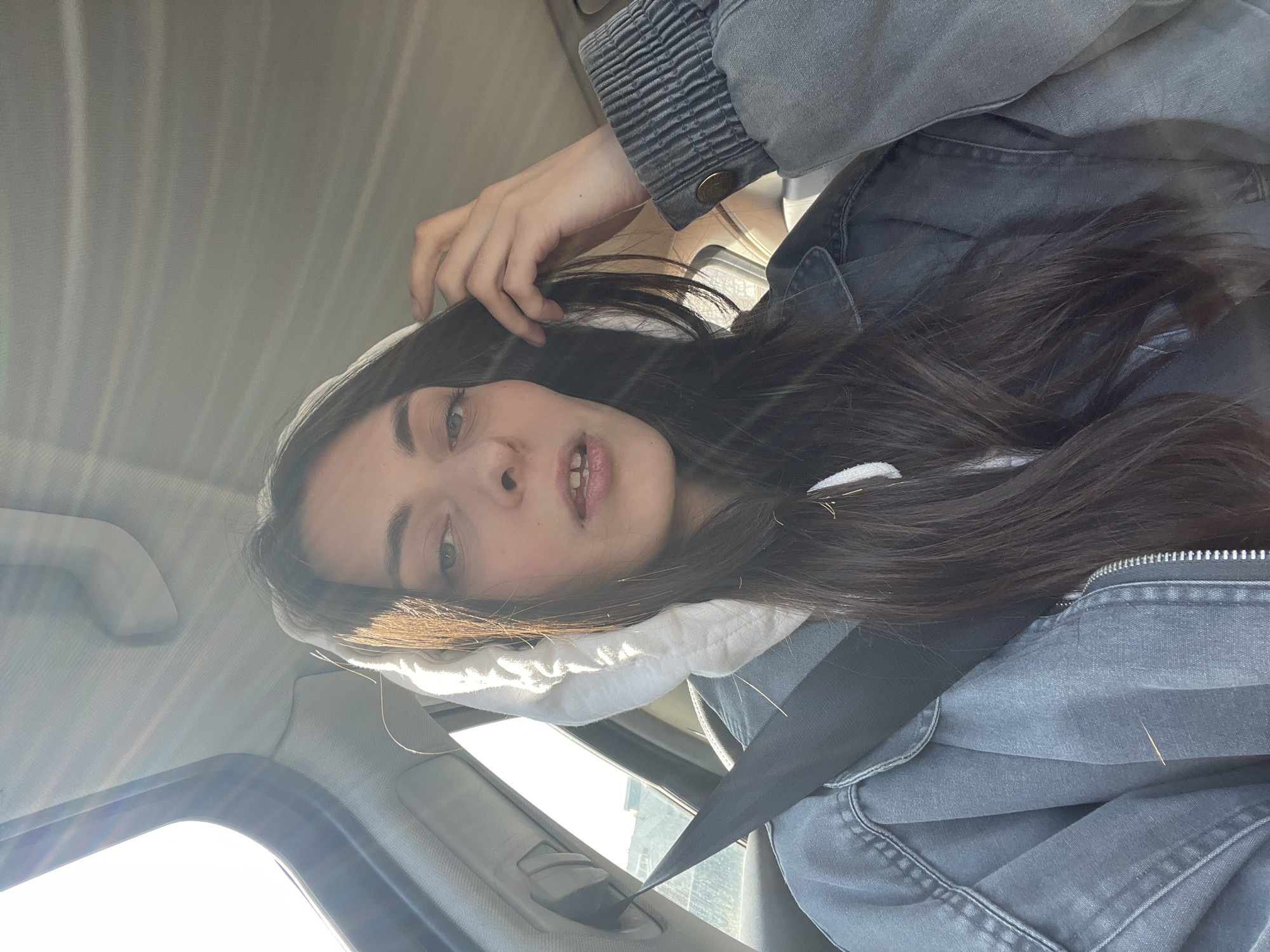 A young person with long, dark hair is captured in a selfie-style photograph taken inside a car. The image is oriented horizontally, with the subject's head towards the right and feet towards the left, implying the original capture was taken vertically and rotated. The subject is looking directly at the camera with a neutral expression, their mouth slightly open, revealing a gap between their front teeth. One hand is visible, gently touching their hair near the top of their head.
They are wearing a grey jacket over a white hoodie, and a black seatbelt crosses their chest, indicating they are either in the driver's or passenger's seat. Bright sunlight streams into the car from the left side of the frame, creating significant lens flare and illuminating parts of the subject's hair and face. The car's interior, including the grey headliner, window frames, and grab handle, is visible in the background, further confirming the setting. The bright light suggests it is daytime and sunny outside. No text is visible in the image.