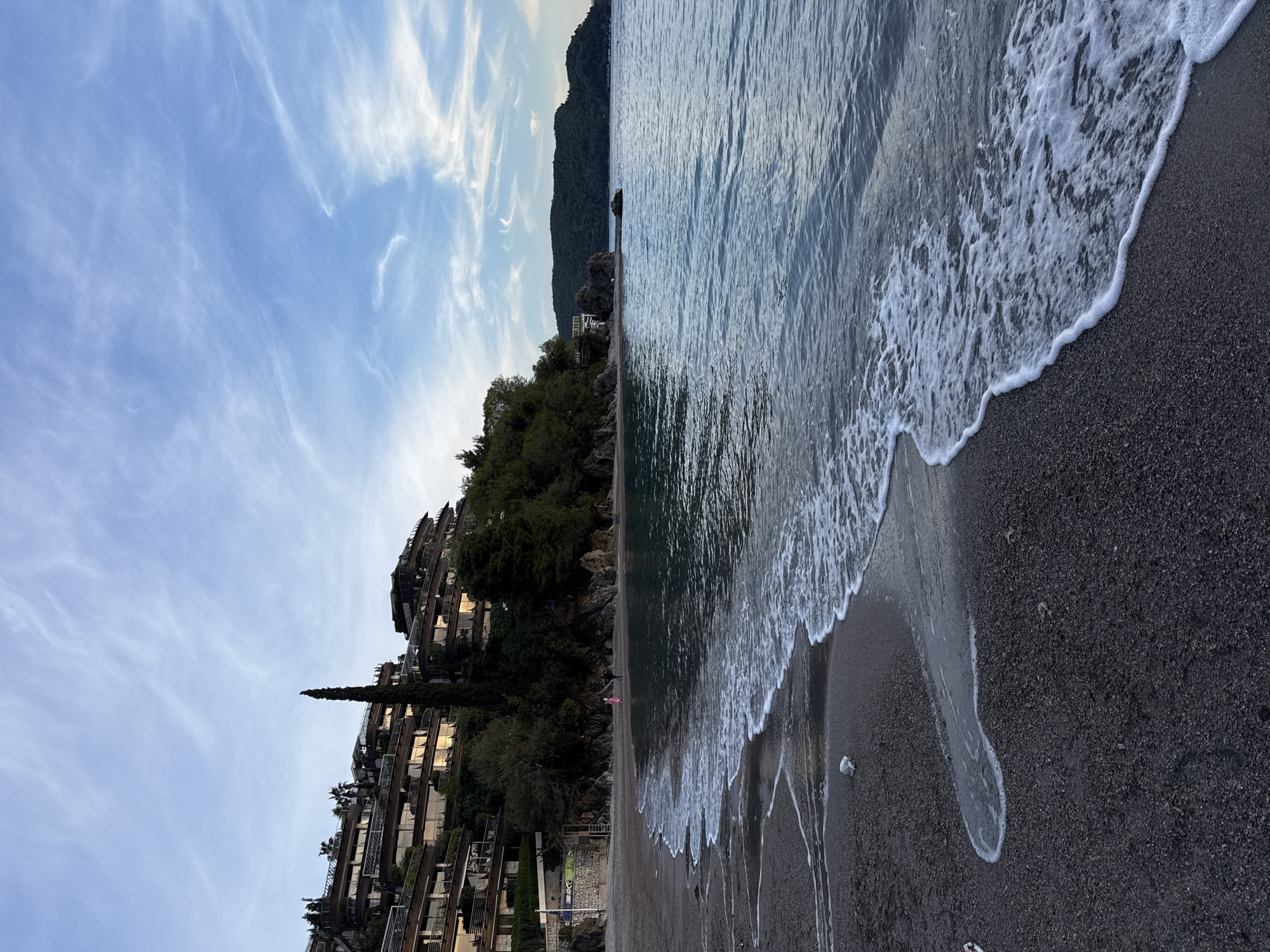 Here is a description of the image:
The image depicts a beach scene, likely during the daytime given the visible sky with scattered clouds. The scene is split between the land on the left and the ocean on the right. On the left side of the image, there is a multi-level building complex situated on a hillside. The buildings appear to be constructed into the natural contours of the land and are surrounded by lush green trees and vegetation.
On the right side, the ocean waves are washing onto a dark-colored sandy beach. The waves are foamy and white as they break onto the shore. There are no visible people swimming or engaging in activities within the ocean.
There is a person standing on the beach.
The location in the city is Unknown, Unknown.