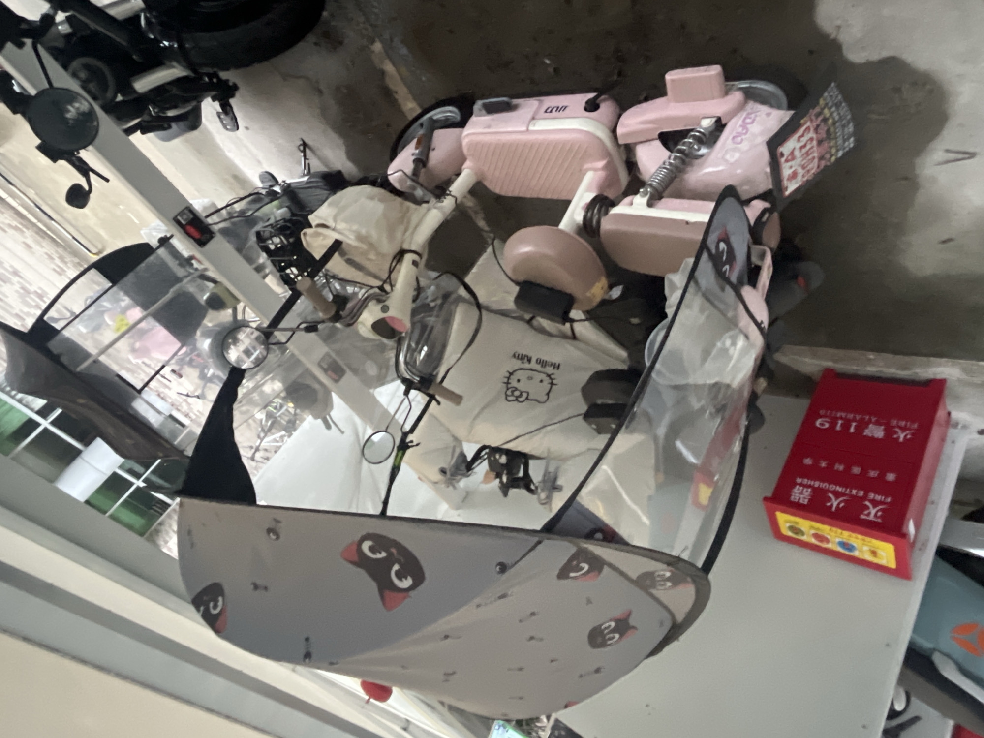 The image displays an indoor or covered parking area within or in proximity to Chongqing Medical University in Yuzhong District, Chongqing, China, as indicated by specific Chinese text on a fire extinguisher box.
In the foreground and central position, a three-wheeled electric scooter is parked. The scooter's chassis is primarily pink and white, equipped with a clear plastic canopy and a grey fabric roof. The fabric roof is patterned with cartoon black cat faces, featuring white eyes and pink inner ears. Circular chrome-rimmed mirrors are mounted on the handlebars, which are partially obscured by a light-colored fabric bag. A white tote bag, branded with the "Hello Kitty" logo, hangs on the front-left side of the scooter. The rear section incorporates a pink, ribbed, rectangular compartment resembling luggage. A black electrical plug or charger is connected to the right side of the scooter's body. A rectangular sign, displaying graphics and text, is attached to the rear, oriented downwards.
To the immediate right of the electric scooter, a portion of a black two-wheeled motorcycle or heavy scooter is visible, characterized by a thick front tire and exposed front suspension.
On the left side of the scene, a red fire extinguisher box with a yellow top panel is mounted on a grey concrete wall. The box is labeled in both Chinese and English, reading "灭火器 FIRE EXTINGUISHER" and "火警 119 FIRE ALARM." Below "FIRE EXTINGUISHER," the characters "重庆医科大学" (Chongqing Medical University) are present. The yellow top panel features four circular indicators or buttons, colored red, yellow, blue, and green.
The floor is a dark concrete or asphalt surface, appearing uneven and stained. In the upper background, a building exterior featuring green-tinted windows framed by grey structural elements is partially visible. No individuals are present in the depicted scene.