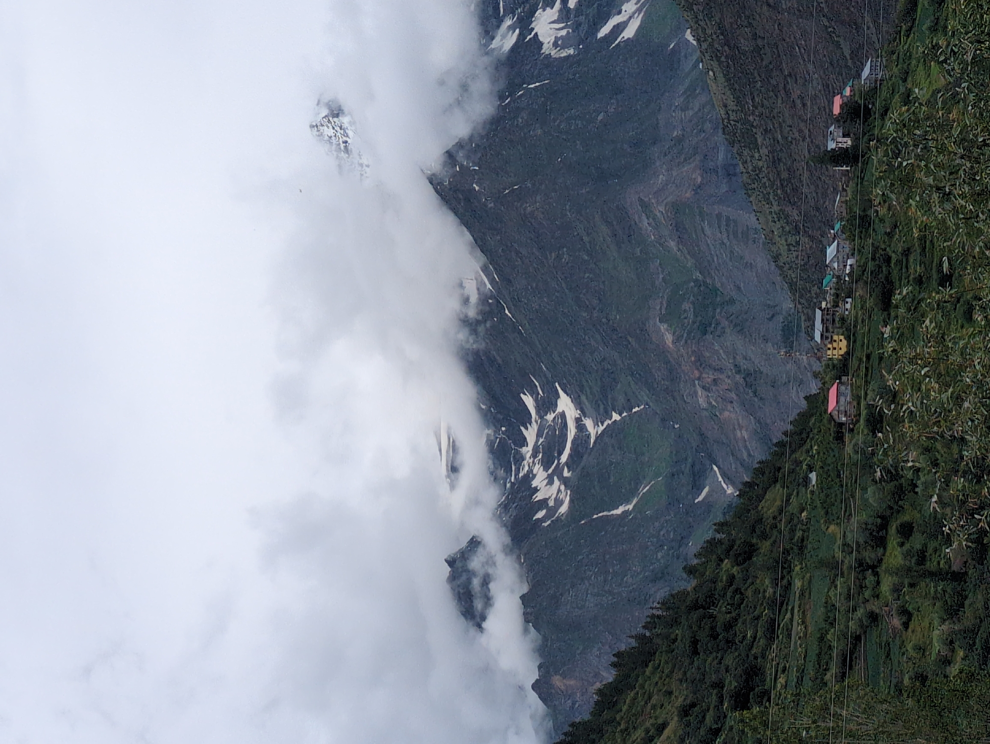 A vertical, medium shot captures a village nestled in a valley of the Himalayan mountains, likely near Keylang, India. The sky is overcast with thick clouds that partially obscure the jagged, dark grey mountain peaks, some of which are dusted with snow. Greenery covers the lower slopes of the mountains, with terraced fields and dark green coniferous trees visible. A small cluster of houses, varying in color and design, are scattered across the hillside. Some houses have red or blue roofs, and one appears to have yellow siding. Power lines run horizontally across the lower portion of the image, partially obscuring the village. The overall mood is somber and slightly dramatic due to the cloudy weather and the imposing mountains.