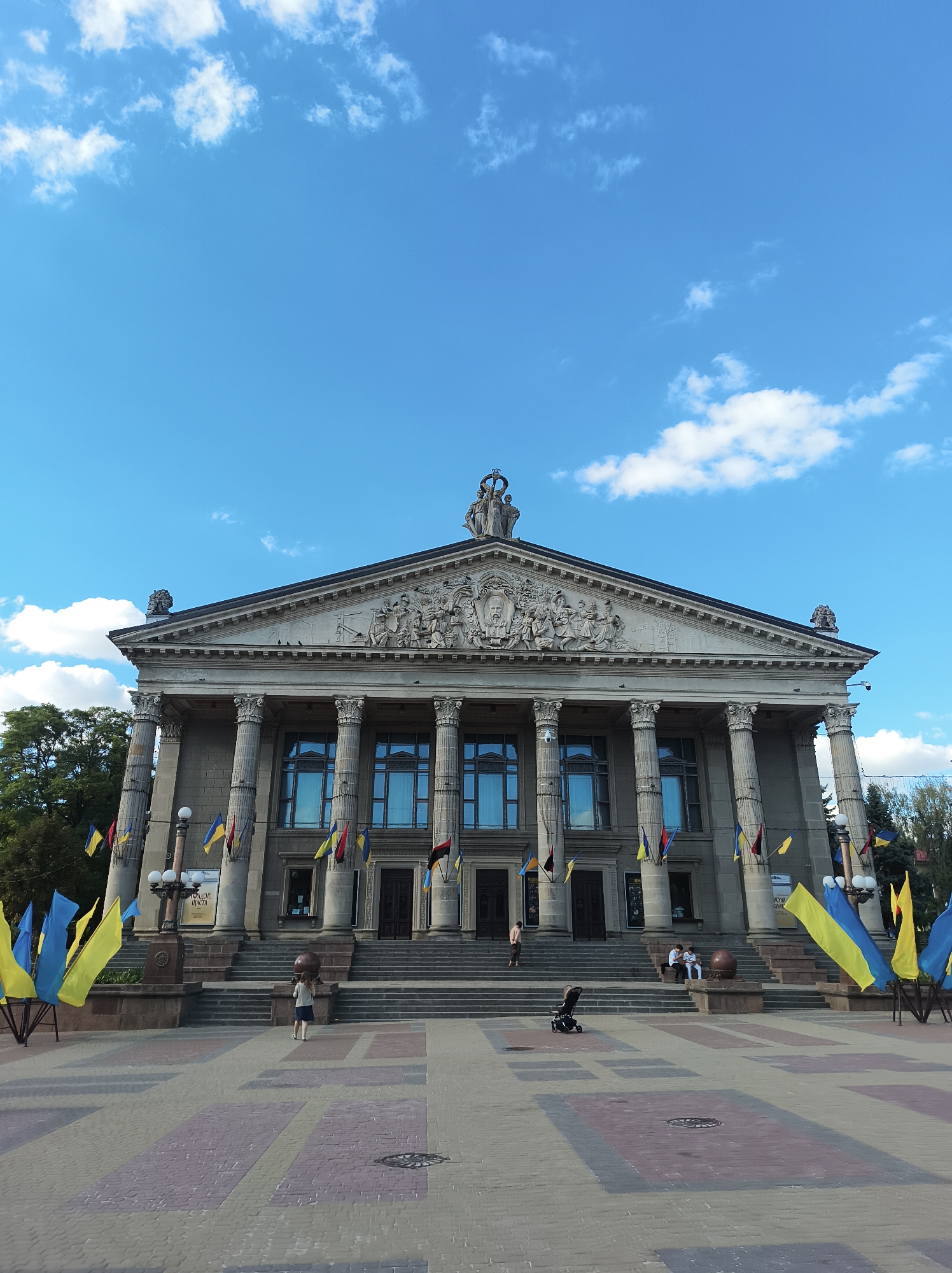 This is a wide-angle, eye-level shot of the Ternopil Regional Drama Theater in Ternopil, Ukraine. The building, a grand neoclassical structure with a prominent portico and columns, is the central subject. It features a decorated pediment with figures and an inscription. Blue and yellow Ukrainian flags are prominently displayed on flagpoles and in front of the building. The foreground is a paved plaza with a geometric pattern of bricks. Several people are visible: a child standing on the steps, two individuals sitting on the steps, and a person walking up the steps on the left side. A stroller is also present in the plaza. The sky is a clear blue with scattered white clouds. Trees are visible behind the theater on the left.