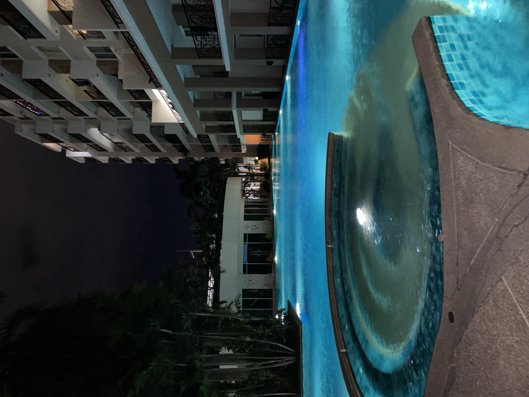 An exterior nighttime photograph captures a swimming pool area adjacent to a multi-story building, likely a hotel or condominium complex, in Pattaya City, Thailand. The pool, illuminated by underwater blue lights, dominates the foreground and mid-ground. A circular, darker pool or jacuzzi with a swirling pattern is situated to the left of the frame, with its edge in the immediate foreground. The main pool extends to the right and into the background, reflecting the ambient light. To the left of the pool, a cluster of tall, slender palm trees stands silhouetted against the dark night sky. A low-rise building with glass doors and windows, identified by a sign reading "POOLSIDE," is visible behind the trees. The building to the right is a tall structure with multiple balconies, some lit, with ornate black metal railings. The overall atmosphere is serene and tranquil, suggesting a resort or residential setting.