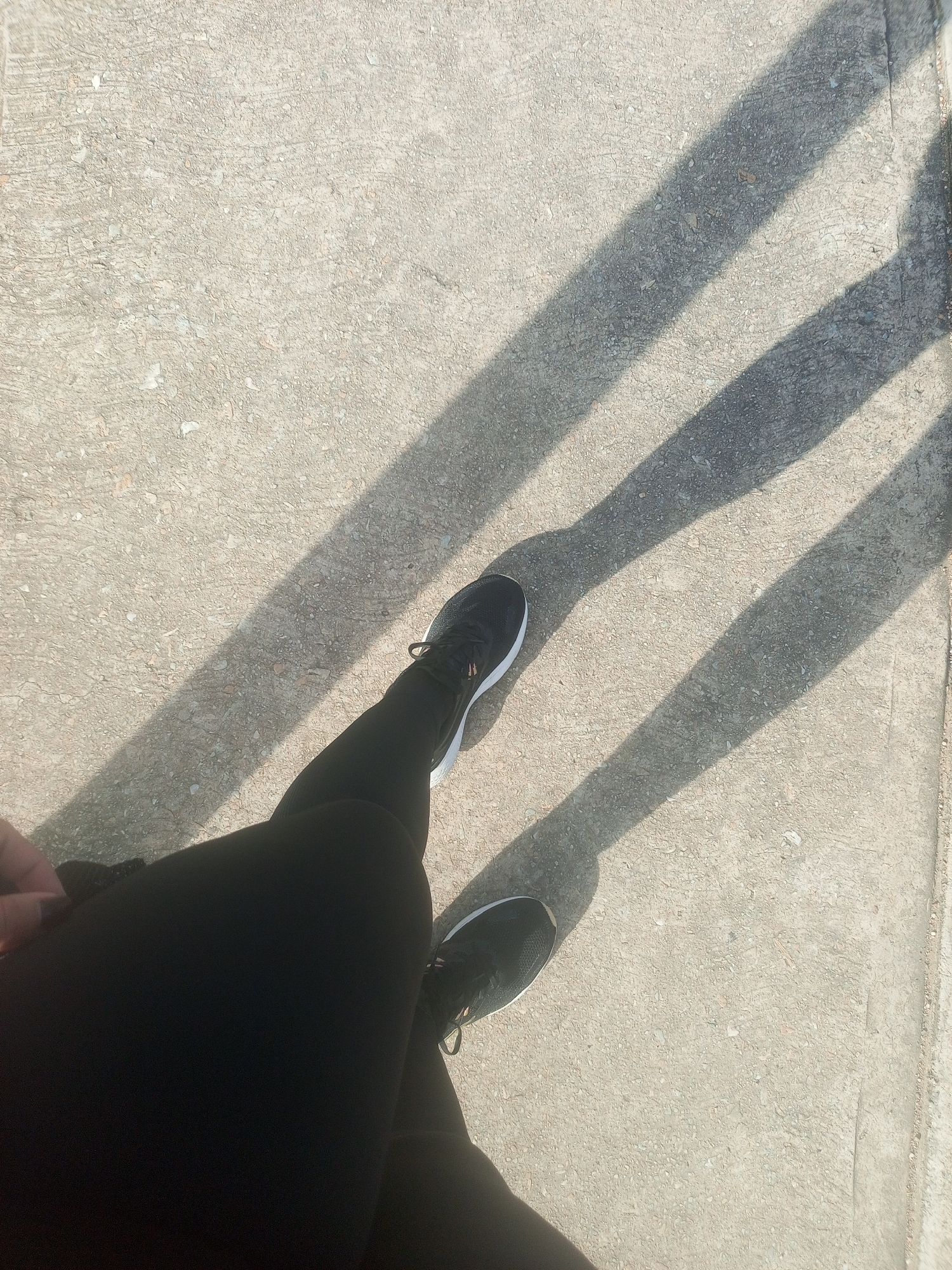 The image depicts a person's legs and feet, clad in black leggings and athletic shoes, standing on a textured concrete surface. Three distinct shadows are cast across the ground, suggesting the presence of overhead structures or objects creating the shadows, possibly a fence or railings. The lighting indicates it is daytime, with strong sunlight. The concrete appears worn and uneven, with small pebbles visible within its composition. The overall scene is minimalist, focusing on the ground and the lower extremities of the individual. There are no other visible people or significant environmental features that would specifically place this scene within Altagracia, Venezuela, or any other urban location.