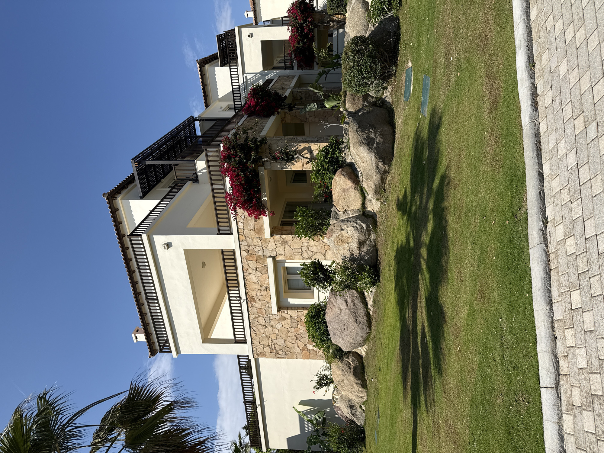 A sunny, outdoor shot displays a multi-story building adorned with flowering vines, nestled alongside a well-manicured lawn, potentially located in Kos, Greece. The building features a cream-colored facade, sections of stone detail, and dark wrought iron balcony railings. Red flowering vines cascade from balconies, adding color to the scene. Landscaping includes rounded boulders, bushes, and green grass. Palm tree fronds are visible on the left edge. A paved walkway borders the lawn. The sky is clear blue, indicative of a bright, cloudless day.