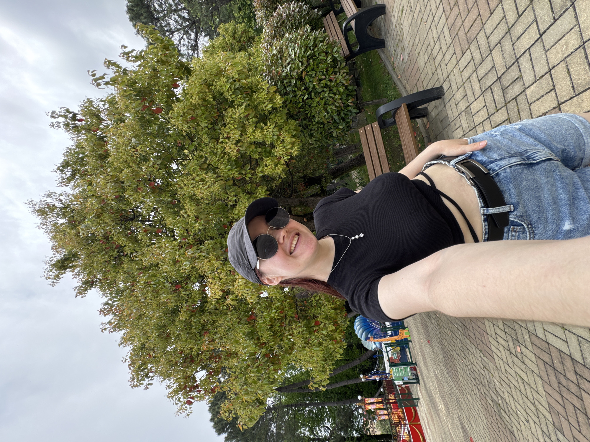 A selfie shows a person sitting on a park bench. The person is wearing a black shirt, denim shorts with a black belt, a gray baseball cap, and sunglasses. They are smiling at the camera and holding the phone with their extended right arm. The park has trees, benches, and a brick walkway. A small amusement park is visible in the background. The city is Sochi, Russia.