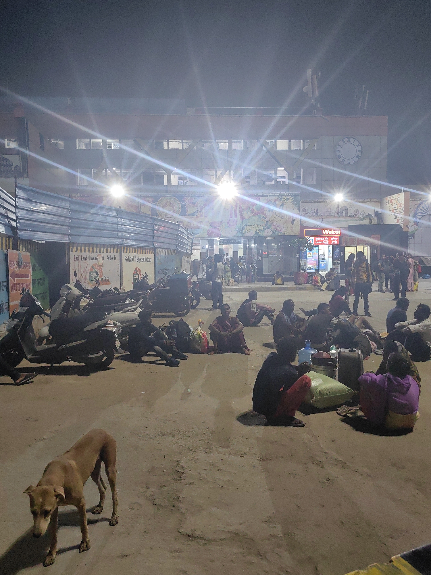 The image depicts a scene at a train station in Delhi, India, at night. A group of people are seen sitting on the ground, many with luggage around them, suggesting they are waiting for a train or have recently arrived. The station building is visible in the background, illuminated by bright lights, with a large clock face on its facade. Several scooters and motorcycles are parked in the foreground, and a stray dog is walking across the dusty ground. The overall atmosphere is one of transient waiting and rest, characteristic of a transportation hub.