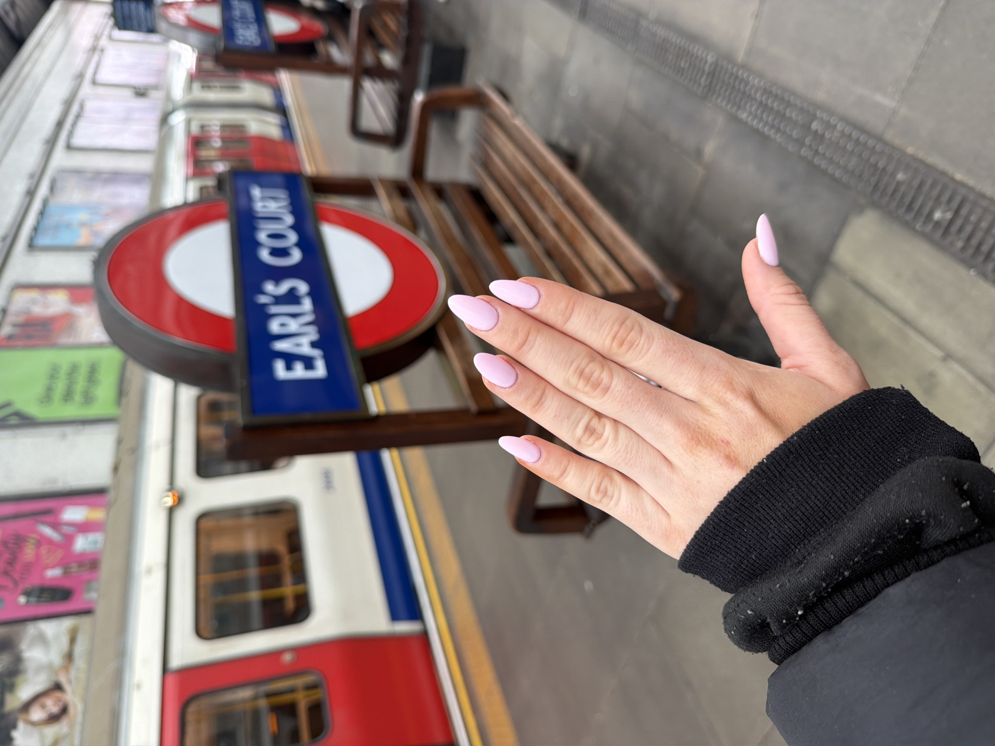 The photograph depicts a woman's hand with light pink almond-shaped manicured nails. She is standing at the Earl's Court Underground station in London, United Kingdom. An Earl's Court station sign is visible, along with a train at the platform. A wooden bench is also in the frame. The woman is wearing a black jacket or coat. The photograph is taken at an angle.