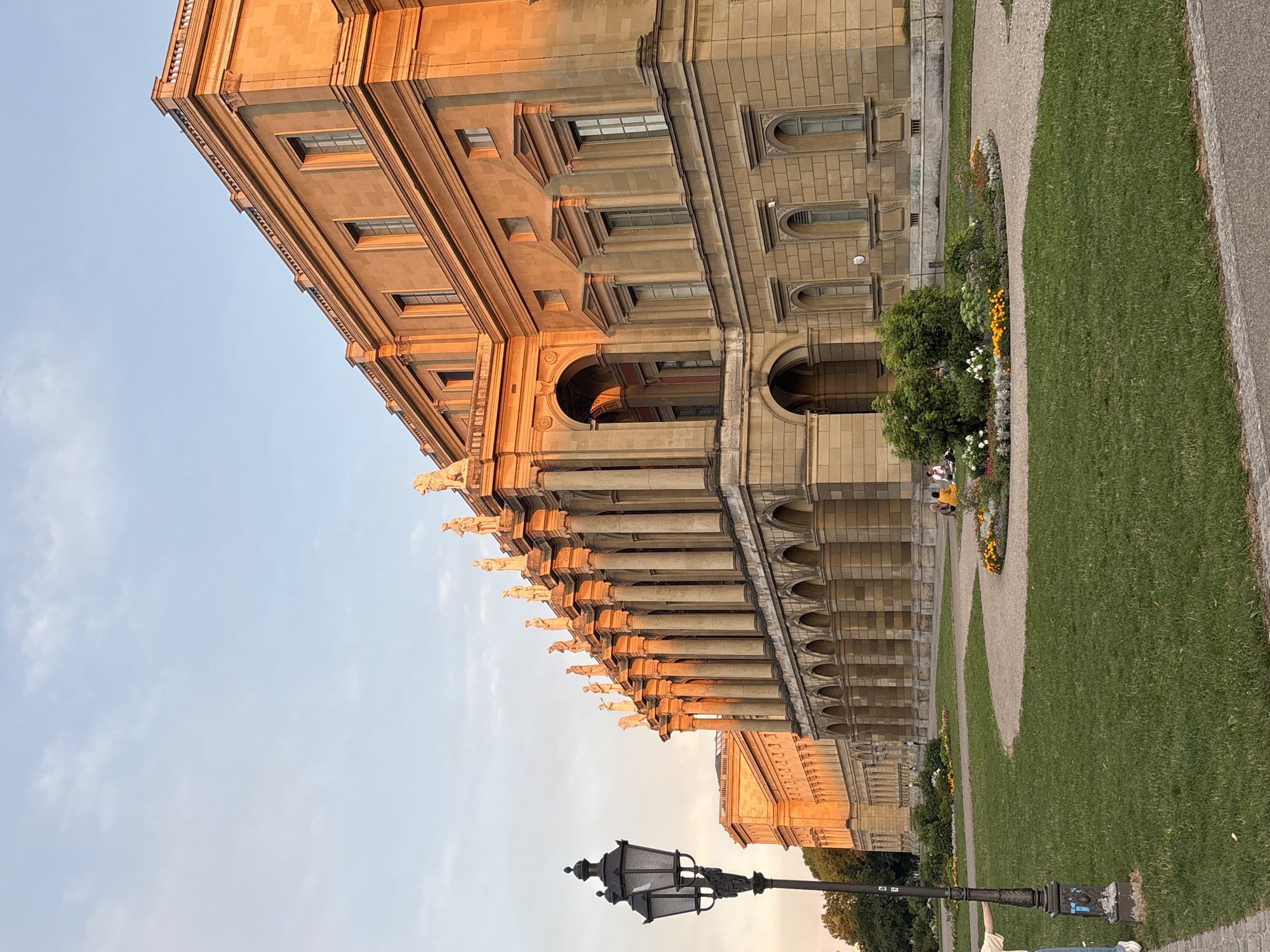 The image captures a view of the Glyptothek museum in Munich, Germany, during a sunset. The warm, golden light of the setting sun illuminates the facade of the classical building, highlighting its colonnades and statues. In the foreground, a well-maintained lawn is divided by a gravel pathway, leading towards the museum. A flower bed with a variety of colorful blooms is situated on the pathway. To the left, a classic black lamppost stands prominently. In the background, a few people are seen relaxing on the grass, and the trees on the left edge of the image suggest a park-like setting surrounding the museum. The sky is a clear, pale blue with a few wispy clouds.