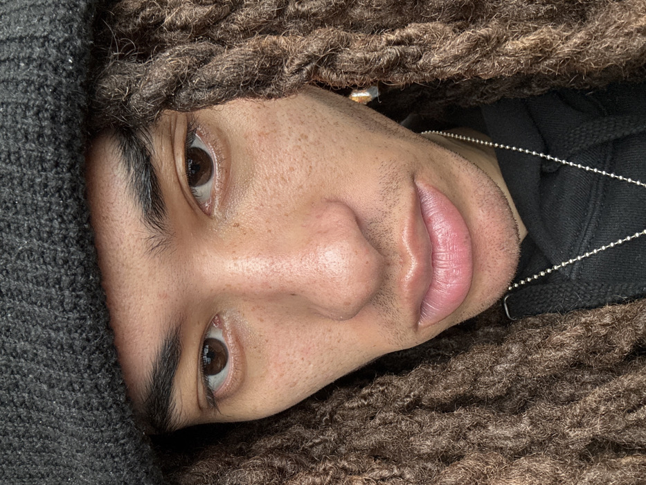 The scene depicts a close-up, head-and-shoulders portrait of an individual, presumably located in the city of Somma Lombardo, Italy. The subject faces directly forward, displaying light brown skin with a notable scattering of freckles across the forehead, nose, and cheeks. Their eyes are dark brown, with thick, dark eyebrows. The nose is centrally prominent, and the lips are full with a light pink hue. Subtle facial stubble is visible on the chin and above the upper lip. The individual wears a dark grey knit beanie that covers the upper portion of the head and forehead. Long, dark brown dreadlocks frame both sides of the face and extend downwards. A small, light-toned, geometric-shaped earring is present in the lobe of the right ear. A thin, silver-colored ball chain necklace is worn around the neck, visible against a black hoodie or sweatshirt worn by the individual. The background is not discernible due to the tight cropping of the image, providing no environmental context.
