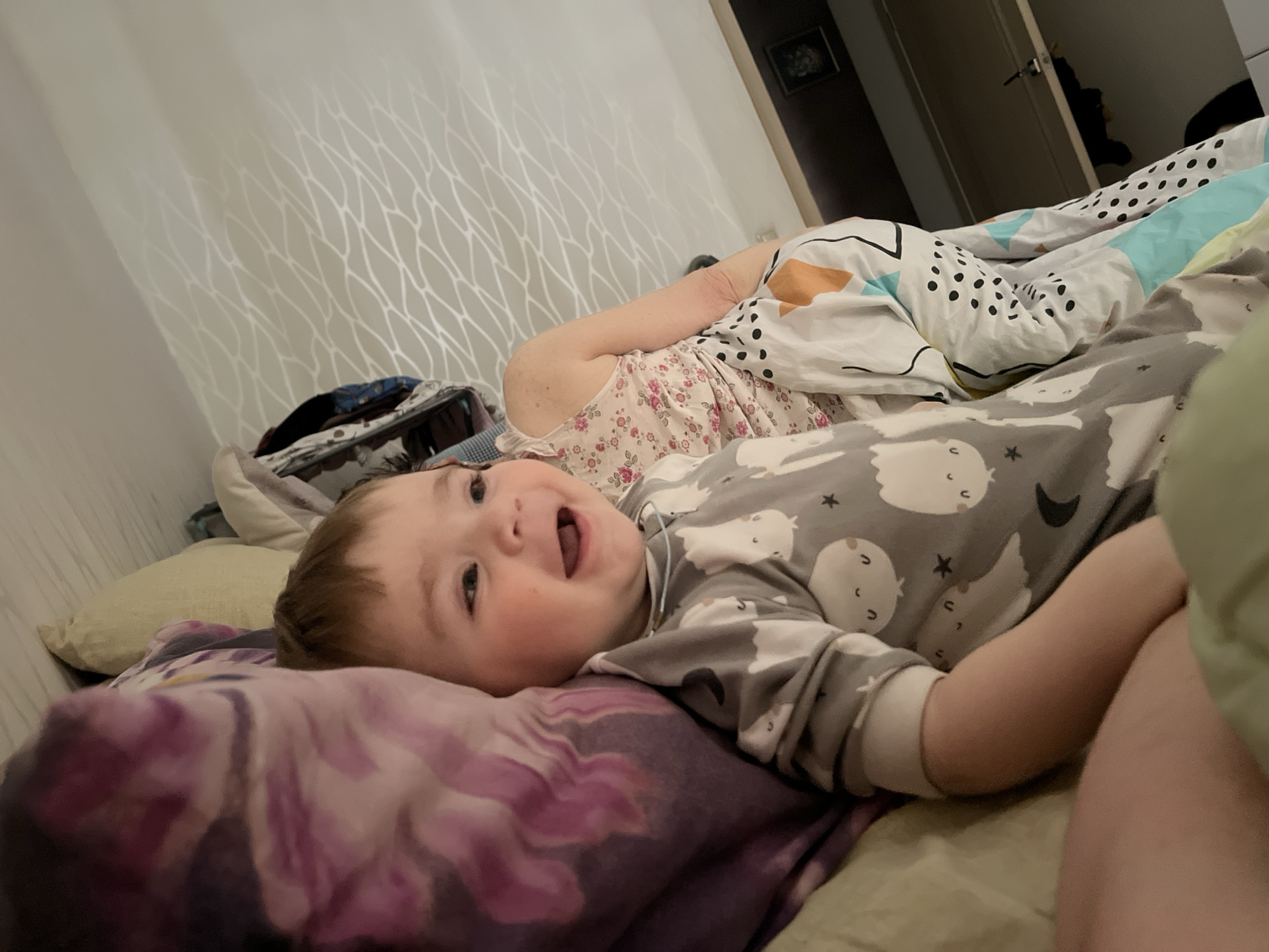 A photograph taken in what appears to be a bedroom, possibly in Perm, Russia, shows a young child, likely under two years of age, lying on a bed with patterned bedding and pillows. The child is wearing a gray pajama top adorned with cartoon images. Their mouth is open, possibly smiling or speaking. An adult is partially visible in the background, also lying on the bed, wearing a floral-patterned top and covered with a patterned duvet. A portion of a door and a framed picture or object on the wall are visible in the background. Luggage can be seen in the background as well. The wallpaper has a wavy pattern. The lighting is artificial, suggesting the photograph was taken indoors.