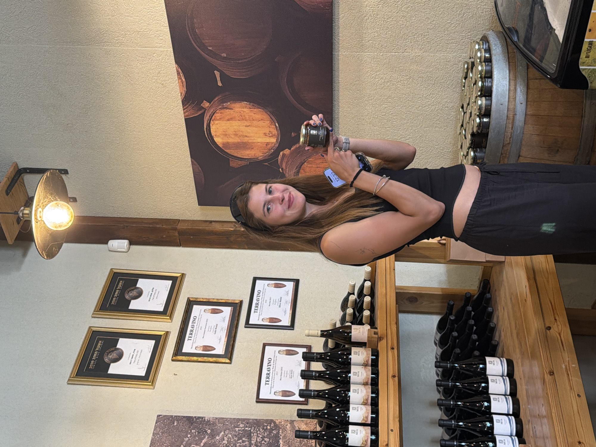 A young woman stands in what appears to be a winery or tasting room, holding a small jar of product and a smartphone. She is wearing a black tank top and dark, loose-fitting pants. She is pointing at the jar she holds in her left hand. Behind her, on the wall, are several framed awards. A shelf below displays numerous bottles of wine. Another shelf to the right holds more wine bottles on their sides, and a large wooden barrel is visible, topped with more jars. The background features a large mural of wine barrels. The scene suggests a commercial establishment, likely a winery, located in the Arava Tichona Regional Council, Israel.
