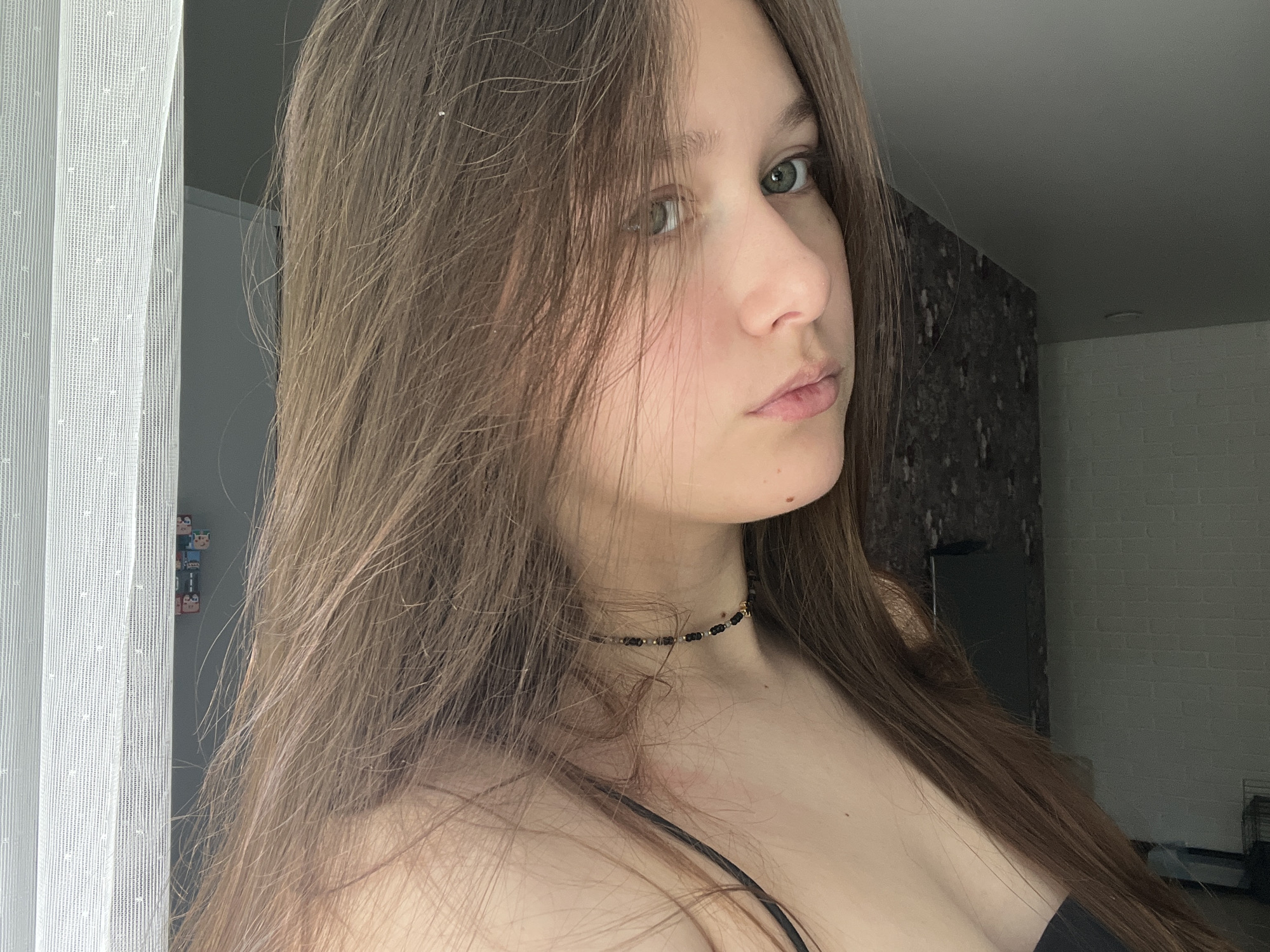 A young woman with long brown hair is posing indoors, looking towards the camera. She has green eyes and is wearing a black beaded choker necklace. She is also wearing a black top with thin straps. In the background, there is a white curtain to the left and brick-patterned wallpaper behind her. The image was likely taken in an apartment or house in Chaikovsky, Russia.