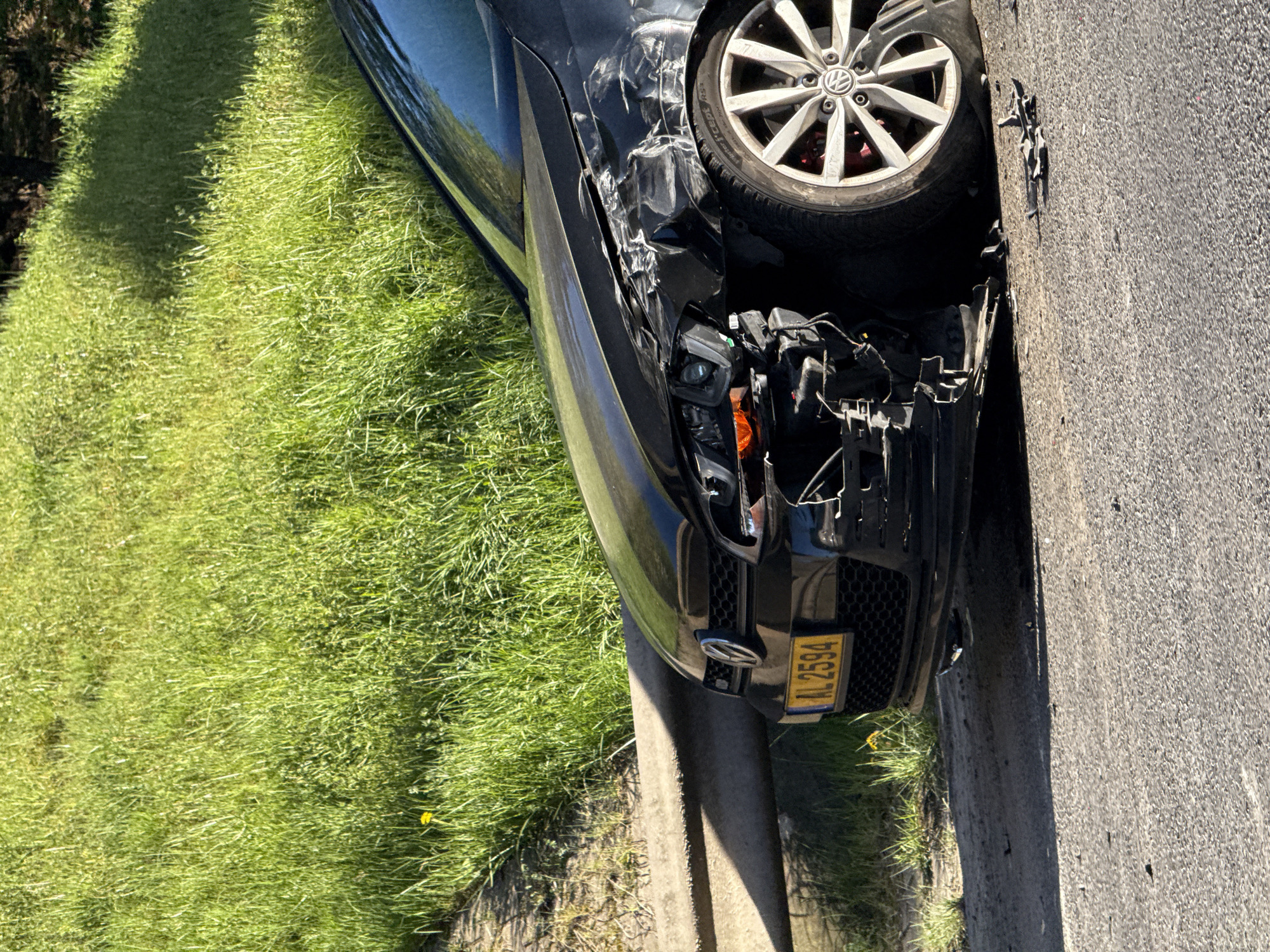 The image depicts a black Volkswagen passenger car involved in a traffic accident. The car's front end, particularly the driver's side, exhibits extensive damage, including a detached bumper, crushed body panels, and exposed mechanical components. The front driver's side wheel is visible. The license plate reads "AL 2594", indicating registration in Luxembourg. The car is positioned on the edge of a road, with a concrete curb separating it from a grassy area. The background includes vegetation and a glimpse of trees. The location appears to be a residential area. The scene suggests a recent collision, based on the extent of the damage. The city is presumed to be Esch-sur-Alzette based on the license plate starting with AL.