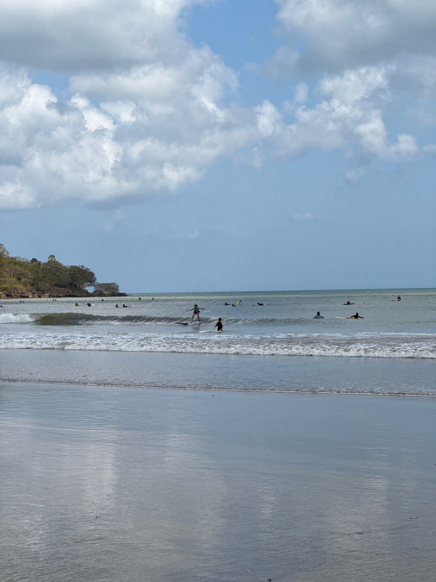 The image depicts a group of surfers in the ocean off the coast of Kuta Selatan, Indonesia. The sea is calm with small waves. Several individuals are on surfboards, some paddling and others standing and riding waves. In the background, a lush, tree-covered hillside forms the coastline. The sky is partly cloudy with a mix of blue sky and white clouds. The foreground shows the wet sand of the beach reflecting the sky and clouds. The scene captures a typical day of surfing at a tropical beach.