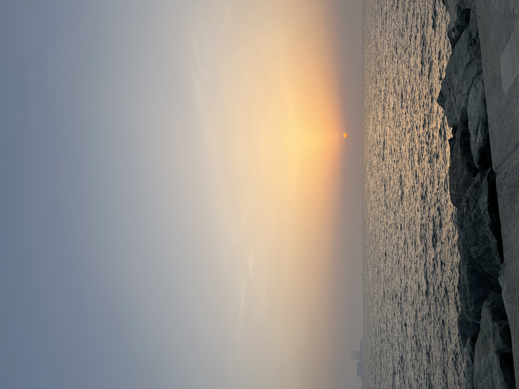 This photograph captures a sunrise or sunset over the water in Dubai, United Arab Emirates. The sun is partially obscured by a thick haze, casting a golden glow across the sky and water. To the right, a rocky coastline or breakwater is visible, composed of large, dark-colored stones. The water is choppy and reflects the golden light of the sky. The sky transitions from a pale blue at the top left to a hazy orange-yellow near the horizon where the sun is positioned. There are a few small, dark spots in the sky that might be birds. The image is oriented vertically.