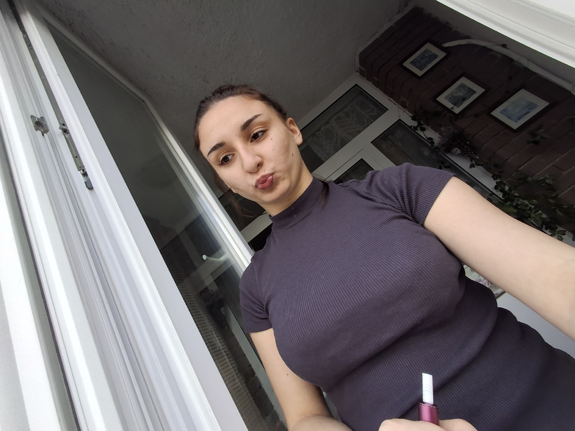 A woman is posing in front of a window. She has brown hair and is wearing a dark purple short-sleeved, high-necked top. She is puckering her lips. In her hand, she holds a small, dark pink object with a white tip. Behind her are windows, a brick wall, and three framed pictures on the wall. The location is not identifiable as Sofia, Bulgaria, from the visible elements in the image.