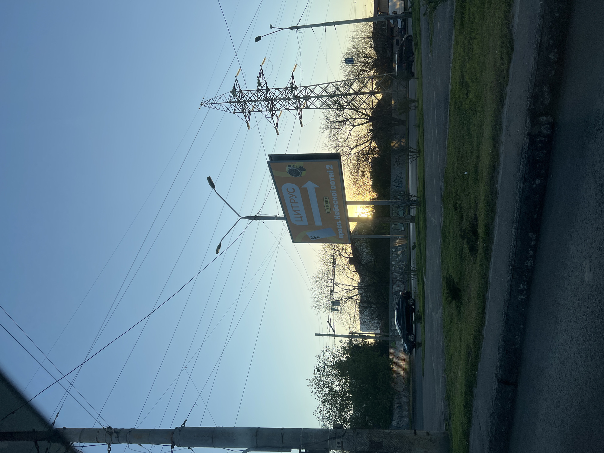 The image depicts a street scene in Odesa, Ukraine, during the late afternoon or early evening, given the position of the sun near the horizon. A large billboard with the Ukrainian word "ЦИТРУС" (Citrus) is prominently displayed. The billboard is advertising "Одесской Сотни 2", which is the address "Odeskoi Sotni 2" street. A high-voltage transmission tower stands tall in the background, with power lines stretching across the sky. The road is flanked by sidewalks and grassy areas. A dark-colored car is parked along the street. The sky is a clear blue with hues of orange and yellow from the setting sun. Street lamps are visible next to the road and the billboard. The overall environment suggests a residential or commercial area on the outskirts of the city.