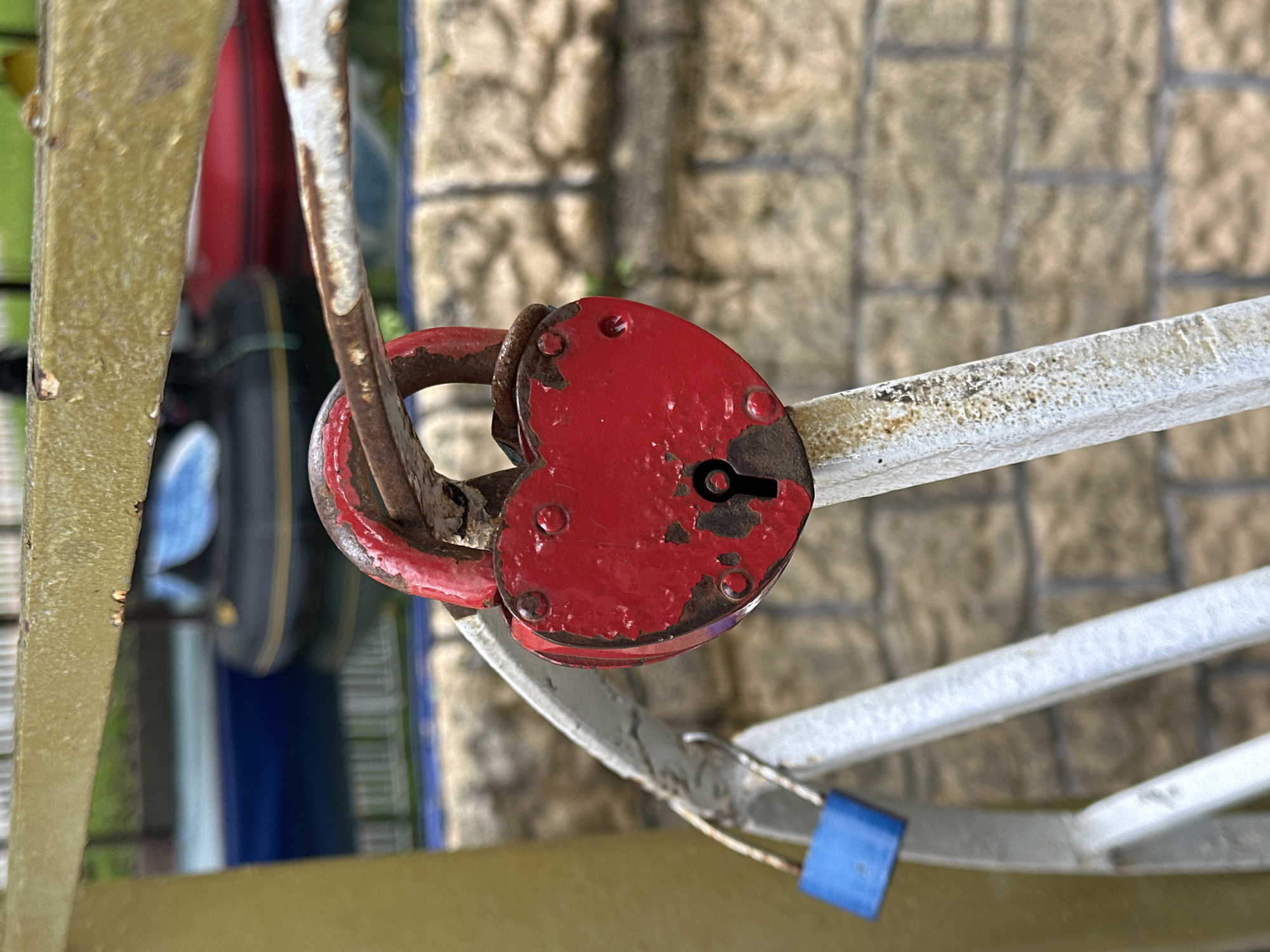 A heart-shaped red padlock secures a rusty metal railing. A small blue padlock is also attached to the railing. The background consists of a brick wall and other blurred structures. Based on the visual evidence, this location is consistent with a "love lock" installation, a common practice worldwide. However, the specific location in Stavropol, Russia, cannot be confirmed without additional information.