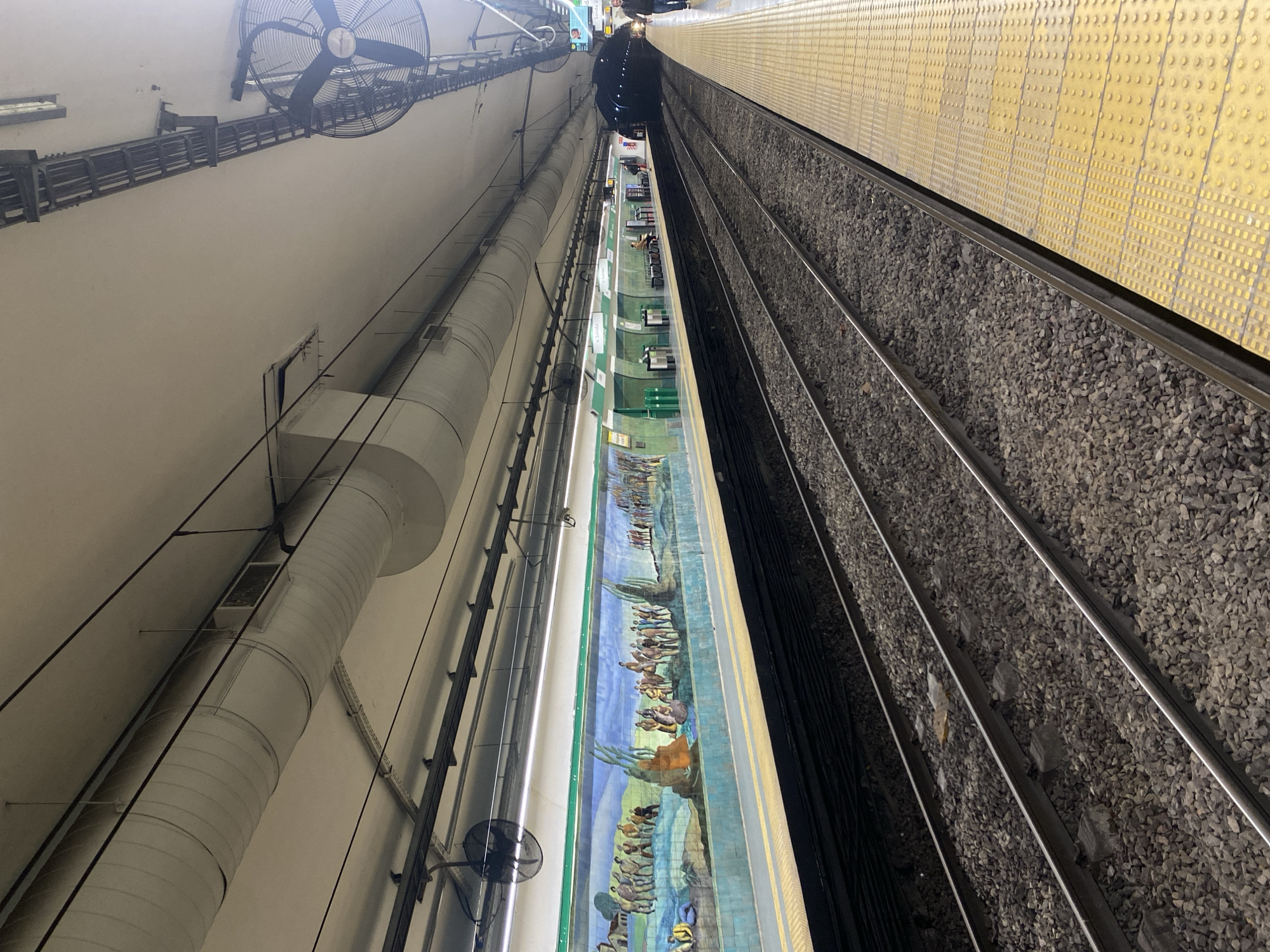 The image shows a subway station in Buenos Aires, Argentina, specifically the Juramento station of the D Line. The view is from an elevated perspective, looking down onto the platform and tracks. The platform features a yellow tactile paving for visually impaired individuals. A mural depicting historical scenes is visible along the platform wall. On the opposite side of the tracks, there is a ventilation system with large ducts and fans. Above, cables and infrastructure run along the ceiling of the station. No people are visible at the moment in the image.
