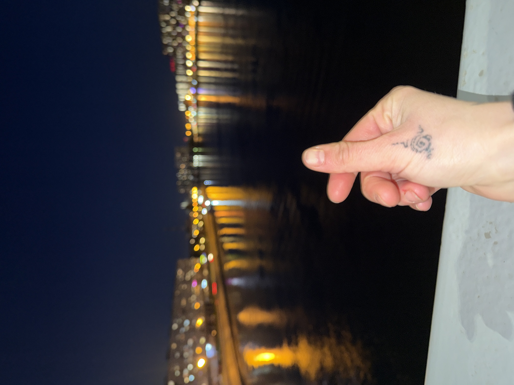 The image displays a nighttime urban scene in Saint Petersburg, Russia, with a prominent focus on a person's right hand in the foreground. The hand is making a "finger heart" gesture, where the thumb and forefinger are crossed to form a small heart shape. A notable detail on the hand is a small, stylized black tattoo resembling a spiral or swirl, located on the side of the palm near the base of the thumb. The hand appears to be resting on or near a light-colored, possibly concrete, railing or wall that runs along the right edge of the frame.
In the background, a body of water, likely a river or canal, stretches across the middle ground, reflecting the blurred lights of buildings on the opposite bank. The city lights appear as vibrant, elongated streaks and circular bokeh effects in shades of yellow, white, orange, blue, and red on the dark water surface, indicating a calm body of water. The sky above is a deep, dark blue, characteristic of a clear night. The activity captured is a casual pose, likely for a photograph, conveying a sense of affection or appreciation for the cityscape. No visible text is present in the image.