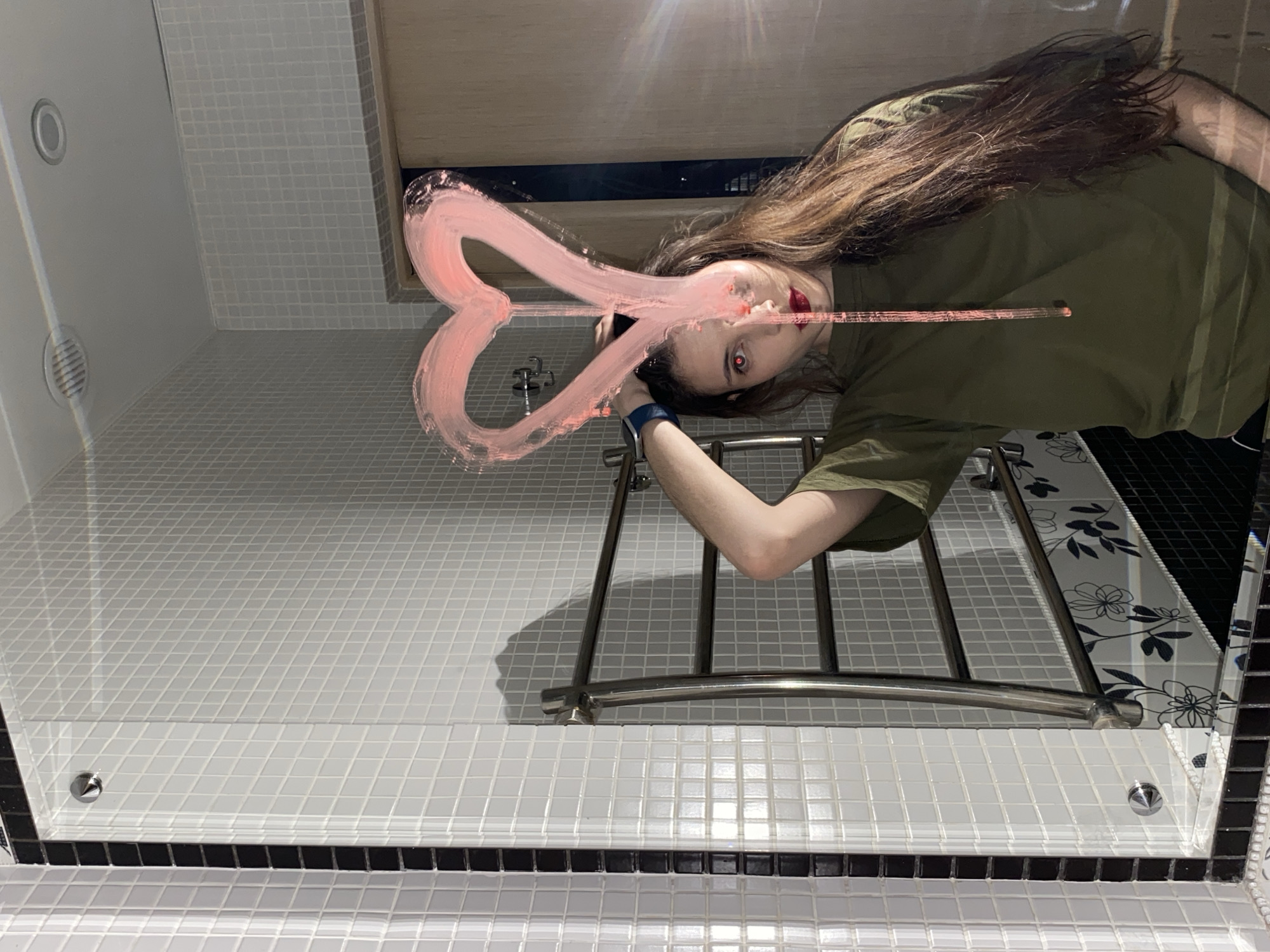 An indoor scene captured from a low angle, looking upwards at a reflective surface, possibly a mirror or glass ceiling, in Votkinsk, Russia. The image itself is rotated, making the reflection appear horizontal.
The primary subject is a young person, reflected upside down on the surface. They have long, dark brown hair and are wearing a plain green t-shirt. Their face is partially obscured by a large, crudely drawn pink heart made of a thick, brush-like substance on the reflective surface. Their lips are painted red, and one eye exhibits a distinct red glow, likely a "red-eye" effect from a camera flash or a strong reflection. The person's right arm is visible, bent at the elbow, suggesting they are holding a device (likely a phone or camera) to capture the image. A dark blue wristband is visible on their right wrist.
The reflected environment appears to be a bathroom, characterized by extensive white square tiles on the walls and floor, with some sections laid diagonally. A chrome metal rack or towel rail is visible below the person's torso. In the upper left of the reflection, circular fixtures, possibly a vent and a recessed light, are visible. A wooden-toned cabinet or shelf is reflected above the person's head. On the far right, black and white tiles with a floral pattern add a decorative element. Mounting screws are visible along the bottom edge of the reflective surface. There is no visible text in the image.