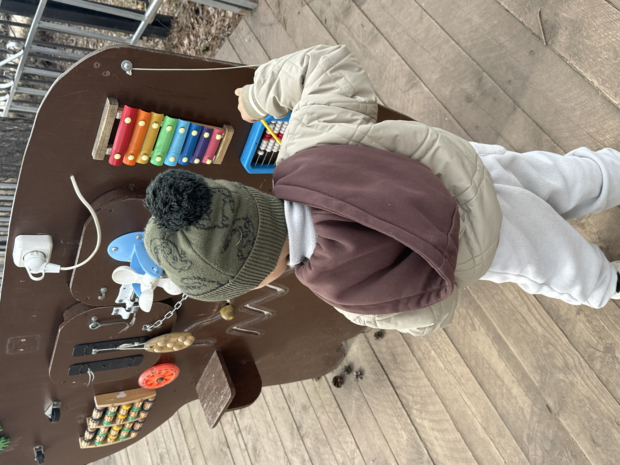 A child wearing a green knit hat with a pom-pom, a brown hooded sweatshirt, a beige quilted jacket, and gray sweatpants is standing at a sensory play board on a wooden deck. The board is brown and has various interactive elements, including a xylophone, an abacus, wooden knobs connected by a chain, gears, latches, a light switch, and a small toy car. The child is holding a yellow and blue tool near the abacus. The setting is outdoors, with stairs visible in the background. The location is Chaikovsky, Russia.