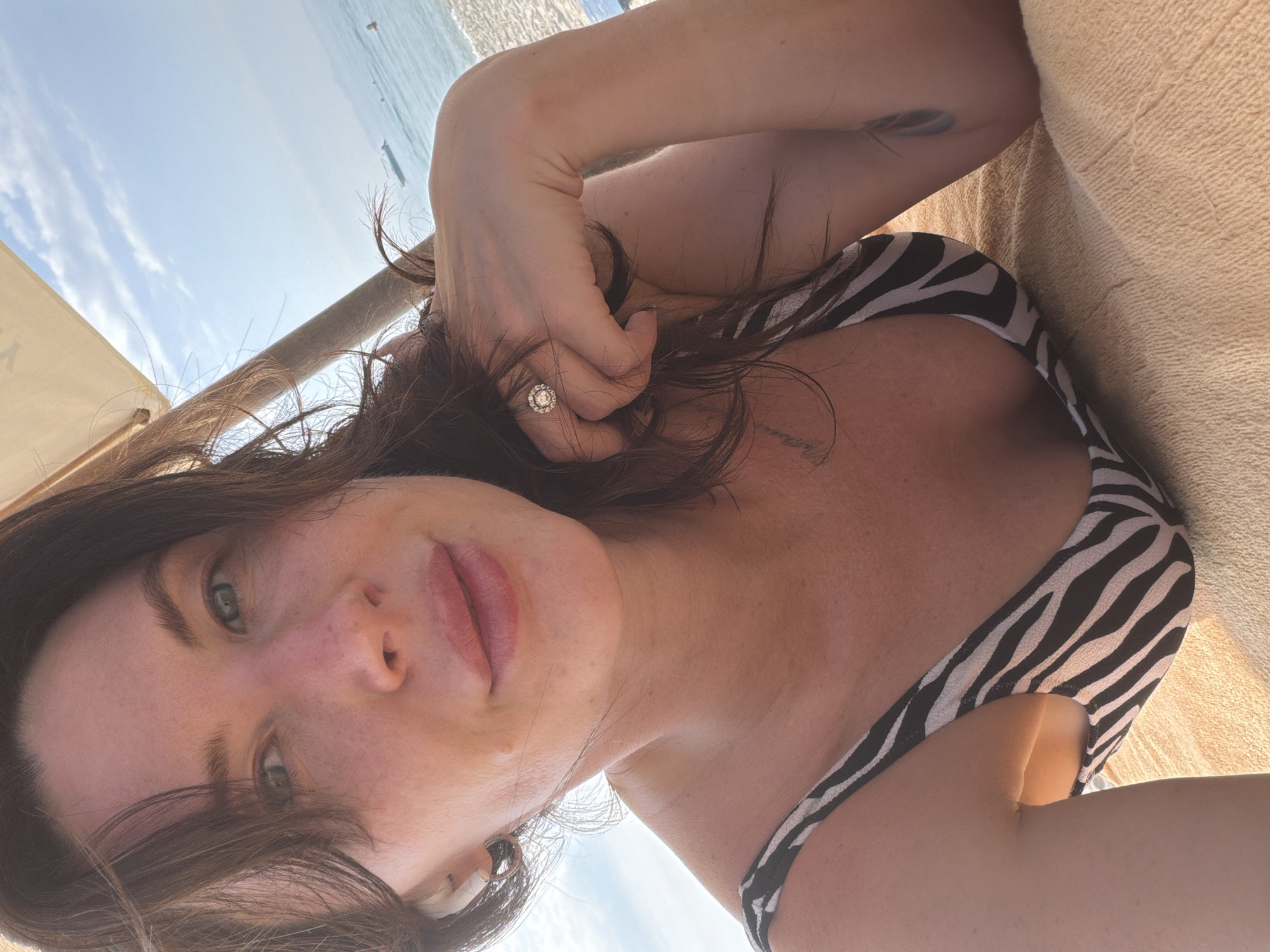 A woman is lying on a beach towel, under a sun umbrella, with the ocean and sky in the background. She is wearing a black and white zebra-print swimsuit. She has dark hair and is wearing a ring on her left hand. The image appears to have been taken in the city of Dong Ninh Hoa Ward, Vietnam, as indicated by the background scenery of a beach with distant boats and a clear blue sky.