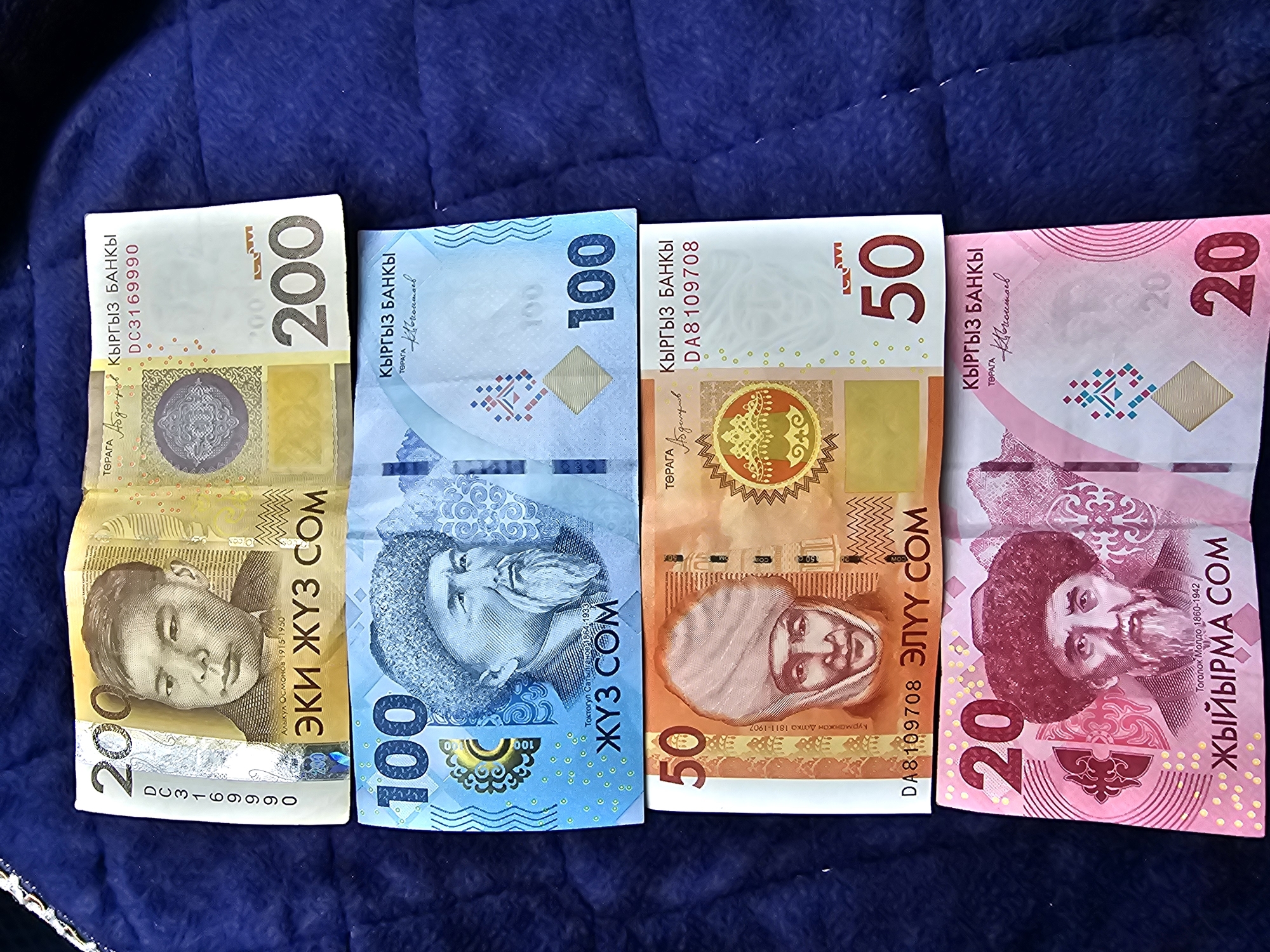 The image displays four Kyrgyzstani Som banknotes, stacked vertically on a dark blue quilted surface, likely a blanket or fabric. The banknotes are from denominations of 200, 100, 50, and 20 Som. The background is out of focus, showing the texture of the fabric. The image is a close-up shot, with the banknotes filling most of the frame. No specific location in the city of Özgön, Kyrgyzstan, can be identified from the image itself, as it only shows the currency.
The top banknote is a 200 Som note, yellow in color, featuring a portrait of a young man on the left and the denomination "200" prominently displayed on the right. The middle section of the banknote has intricate patterns and text in Kyrgyz.
Below the 200 Som note is a 100 Som note, predominantly blue. It features a portrait of an elderly man with a beard on the left and the denomination "100" on the right. This note also has decorative elements and Kyrgyz text.
The third banknote from the top is a 50 Som note, with orange and beige tones. It depicts a portrait of a person wearing a distinctive headscarf on the left and the denomination "50" on the right.
The bottom banknote is a 20 Som note, in shades of pink and purple. It shows a portrait of a man with a beard and a traditional hat on the left and the denomination "20" on the right.
The banknotes appear to be in good condition, with crisp folds visible on some of them. The lighting suggests an indoor setting or diffused natural light.