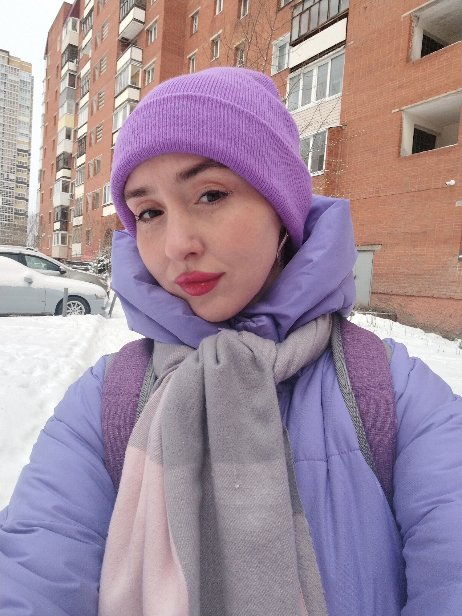 The image displays a close-up selfie of a young woman taken outdoors in a snowy urban environment, identified as Izhevsk, Russia. The woman is the primary subject, centered in the frame. She wears a vibrant purple knitted beanie, a light lavender puffer jacket with a high, protective collar, and a thick grey and pink plaid scarf wrapped around her neck. The straps of a purple backpack are visible over her shoulders. She has fair skin, dark eyes with visible eyelashes, defined eyebrows, and bright red lipstick, looking directly at the camera with a neutral to slightly serious expression.
The background reveals a typical residential area in winter. To the left, a tall, modern, high-rise building is partially visible, alongside a silver car heavily covered in snow. To the right and center, a large, multi-story brick apartment building with numerous windows and balconies dominates the background. The ground is entirely blanketed in deep white snow, indicating cold, winter weather. The sky appears overcast, suggesting daytime. No discernible text is visible within the image.