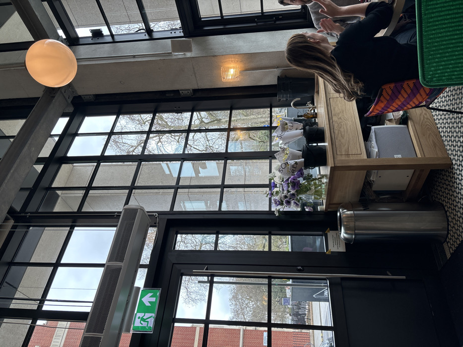 The image depicts the interior of a cafe or restaurant located on Arundel Street in the City of Westminster, London, United Kingdom. The scene is dominated by tall, black-framed industrial-style windows that span much of the wall, providing a view of an urban street with trees and a red brick building. Above a black-framed glass door on the left, a green emergency exit sign with a running person and an arrow pointing right is visible, along with a long, metallic air conditioning or heating unit.
In the mid-ground, a light wooden counter or island features a sink with a faucet. On this counter, a clear glass vase holds purple and white flowers, and several bouquets wrapped in white paper are displayed in black buckets. A dark bottle and a white electronic device, possibly a speaker, are also present on or below the counter. A tall, stainless steel cylindrical bin stands next to the door.
To the right, two individuals are seated at a table. One person, seen from the back and side, has long brown hair and wears a dark top. They are seated in a chair with an orange and purple geometric pattern and are actively gesturing with their hands during a conversation. Part of another person, with visible facial hair, is seated opposite them, partially obscured on the far right.
Overhead, a large, spherical, illuminated pendant light hangs from the ceiling. A modern floor lamp with a woven-patterned shade casts a warm glow from near a concrete pillar on the right. The floor exhibits a patterned black and white tiled design. Natural light largely illuminates the space, supplemented by the artificial light sources.