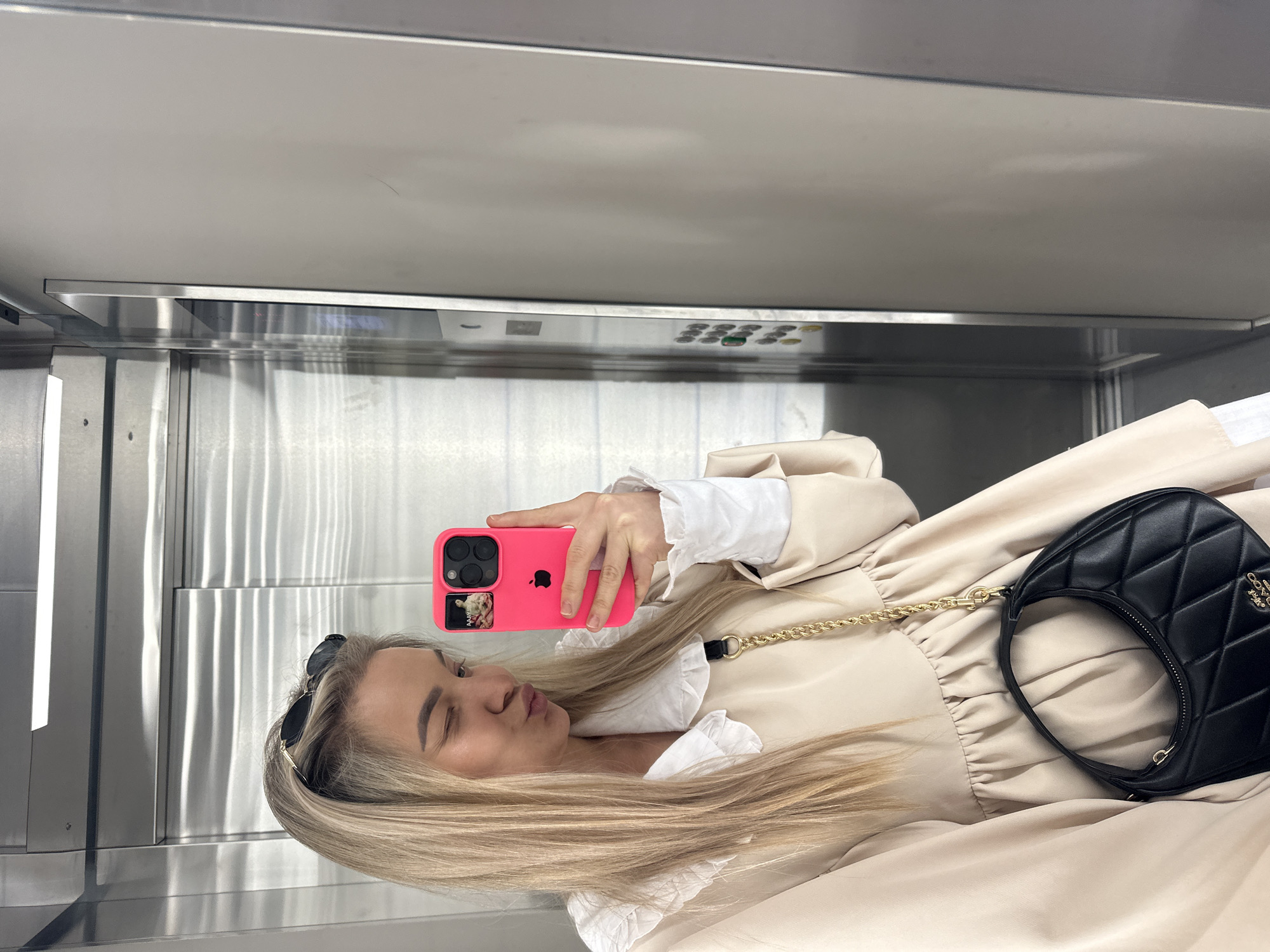 A blonde woman is taking a selfie in what appears to be an elevator. She holds a pink phone with a decorative sticker on the back. She is wearing a light beige dress with white ruffled details at the collar and cuffs. A black, quilted handbag with a gold chain strap is visible. Sunglasses are perched on top of her head. The elevator interior is stainless steel, and a control panel is visible above her. The city of Budva, Montenegro, is not discernible from the image.