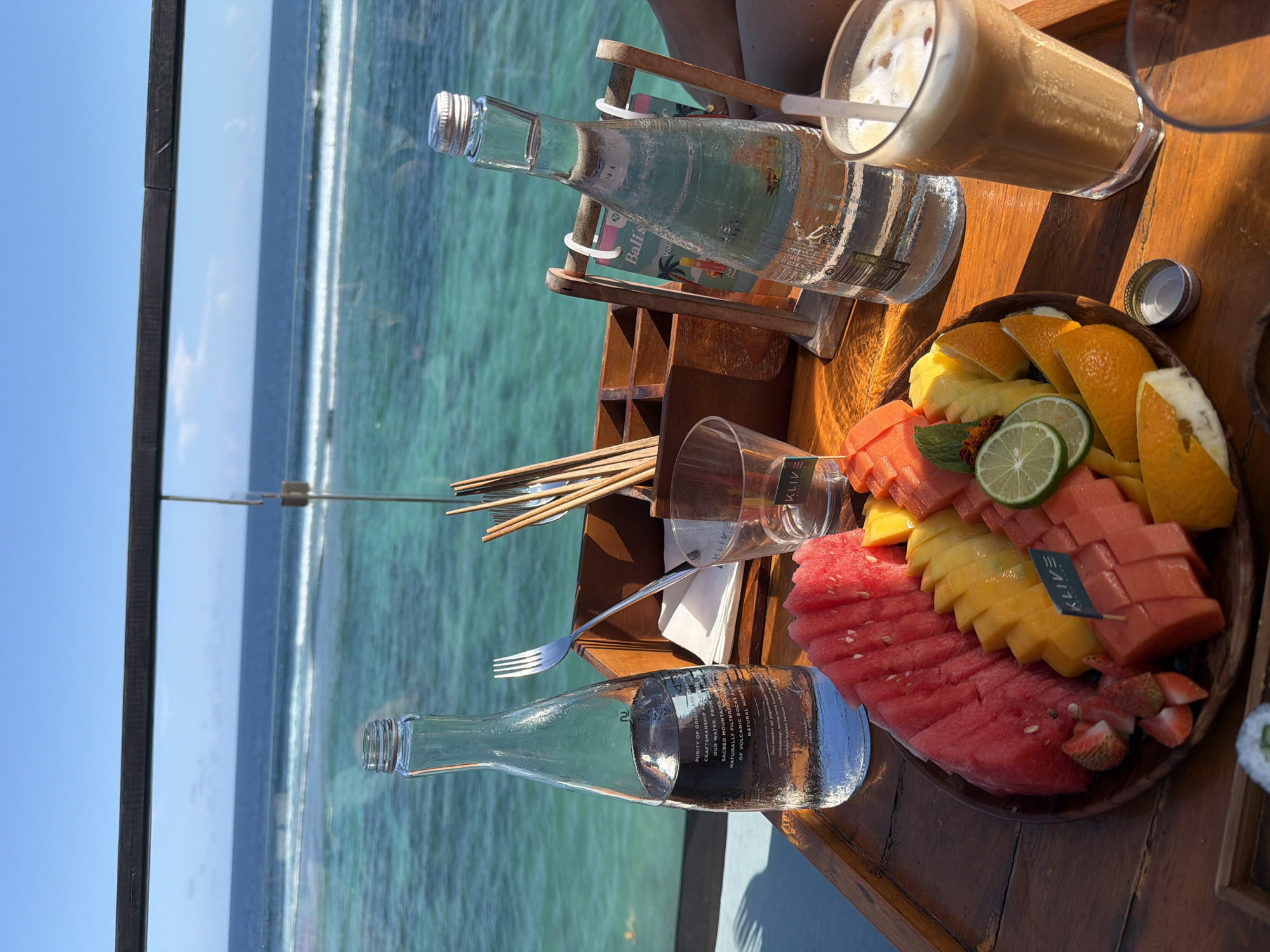 Here's a detailed description of the image:
**Scene:** A close-up, outdoor dining setting with a view of the ocean. The image is taken from a high angle, looking down onto a wooden table.
**Location:** Possibly Ungasan, Indonesia (based on text visible on one bottle).
**Objects:**
* **Food and Drink:** A fruit platter featuring watermelon, mango, oranges, papaya and lime slices. A glass of iced coffee with a straw, two glass bottles of mineral water, and a small dish with sushi (partially visible).
* **Table Setting:** A wooden serving tray with individual compartments holding wooden skewers, a fork, and a drinking glass.
* **View:** The ocean is visible in the background, along with the sky. The water appears turquoise and there are distant waves.
**Environment:**
* The setting appears to be a restaurant or a deck overlooking the sea.
* The lighting suggests it is daytime.
* The overall scene suggests a relaxing, vacation-like atmosphere.
**Actions/Interactions:** N/A, the scene is static.