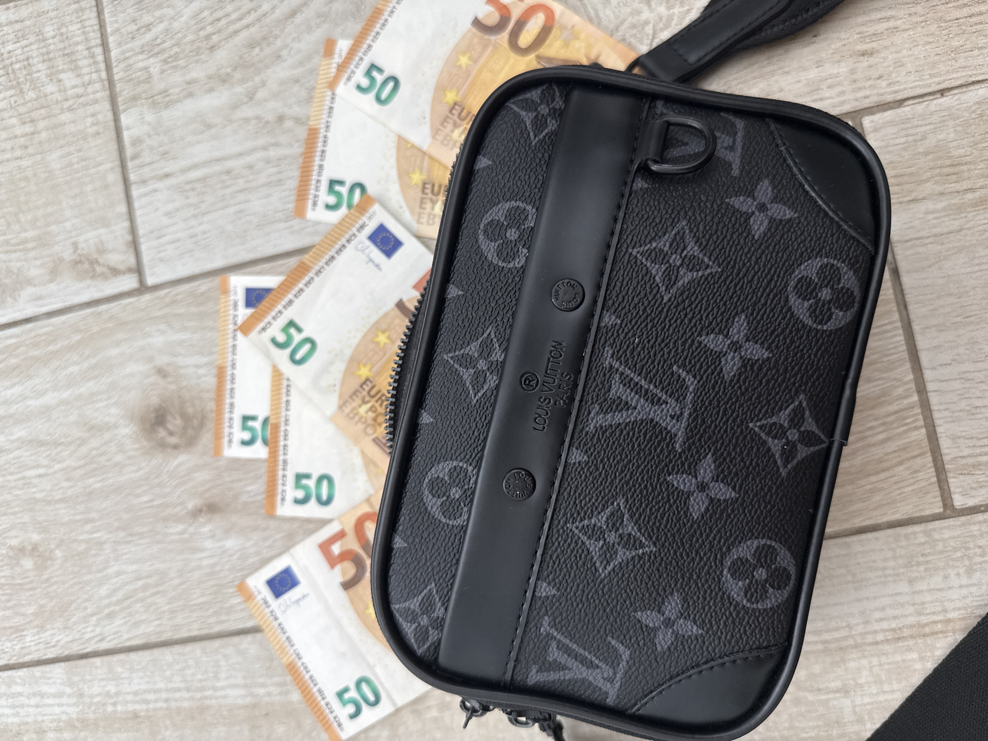 The image shows a close-up, overhead view of a black Louis Vuitton crossbody bag, patterned with the brand's signature monogram. The bag is placed on a light-colored wooden surface, which appears to be a floor or tabletop. Several euro banknotes, specifically 50 euro notes, are scattered around and beneath the bag. The bag is oriented horizontally, with the front facing the viewer. The details of the bag's texture, stitching, and metal hardware are clearly visible. The banknotes are slightly creased and overlapping, suggesting they have been handled. The overall lighting is bright and even, indicating it was taken indoors or in diffused daylight. There is no indication of the city of Burgas, Bulgaria, in the image or its context.