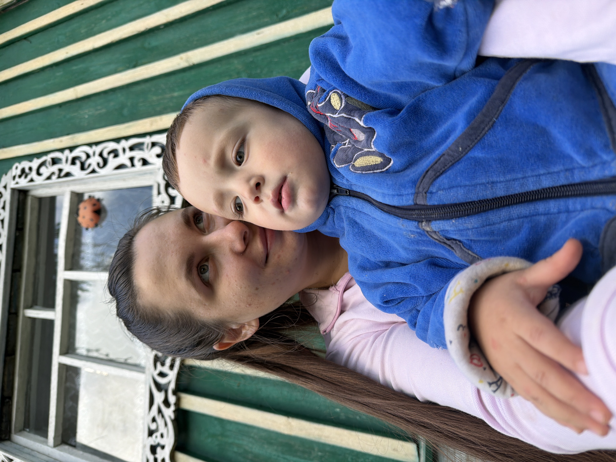 In Красный Маяк, Russia, a woman with long brown hair tied in a ponytail and wearing a pink shirt holds a young child. The child is wearing a blue hooded jacket with a zipper and a patch on the chest. They are positioned in front of a green wooden wall with white trim and a window. A decorative element resembling a ladybug is visible in the window. The scene appears to be outdoors, possibly near a residential building.