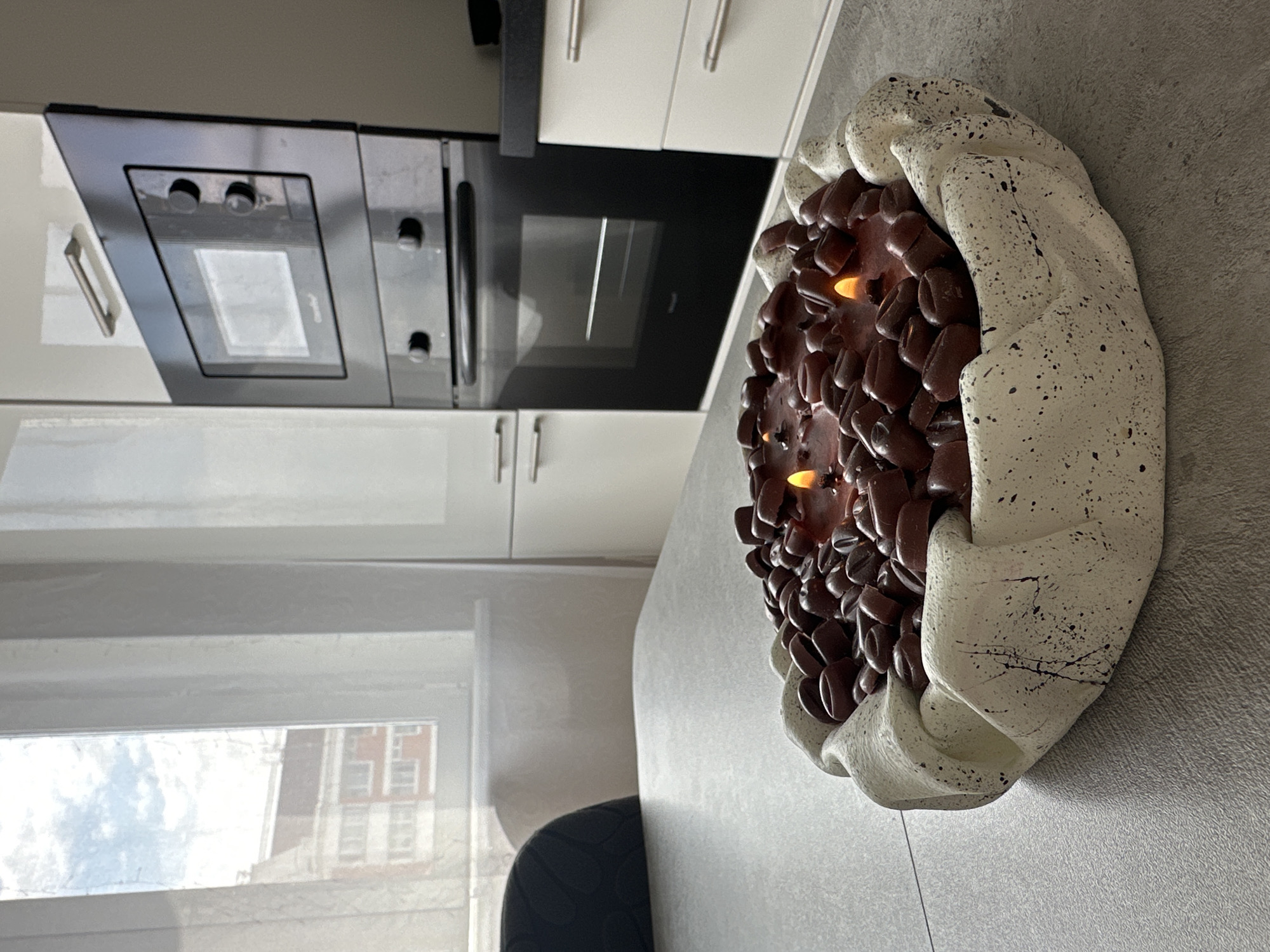 The image depicts an indoor scene, likely a kitchen, with a large, decorative candle in the foreground. The candle is housed in a rough-textured, off-white bowl and is filled with dark brown, bean-shaped objects, possibly resembling coffee beans, embedded in a reddish-brown wax. Two small flames are visible, indicating the candle is lit.
Behind the candle, a grey countertop is visible, leading to kitchen cabinetry. To the left, a portion of a dark upholstered chair is present. In the background, a window with sheer white curtains reveals a glimpse of an apartment building with red brick accents and a cloudy sky. Adjacent to the window, a modern black built-in oven and microwave unit is installed in white cabinetry. The overall environment appears to be a domestic setting. The image does not provide any information about the city of Malye Ust'ya, Russia.