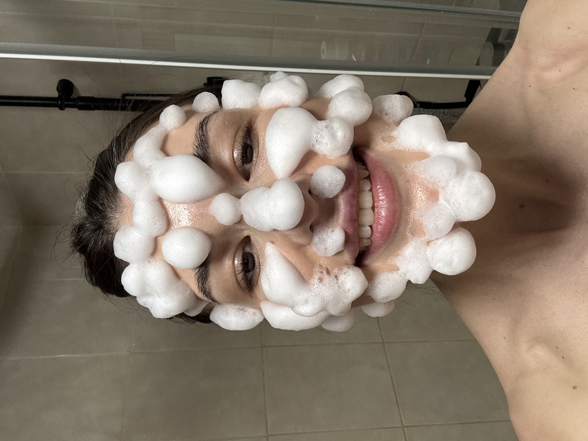A woman with dark hair has soap bubbles covering her face. She is smiling, revealing her teeth. She appears to be in a bathroom, with tiled walls and a shower fixture visible in the background. The location in Chaikovsky, Russia, cannot be determined from the image.